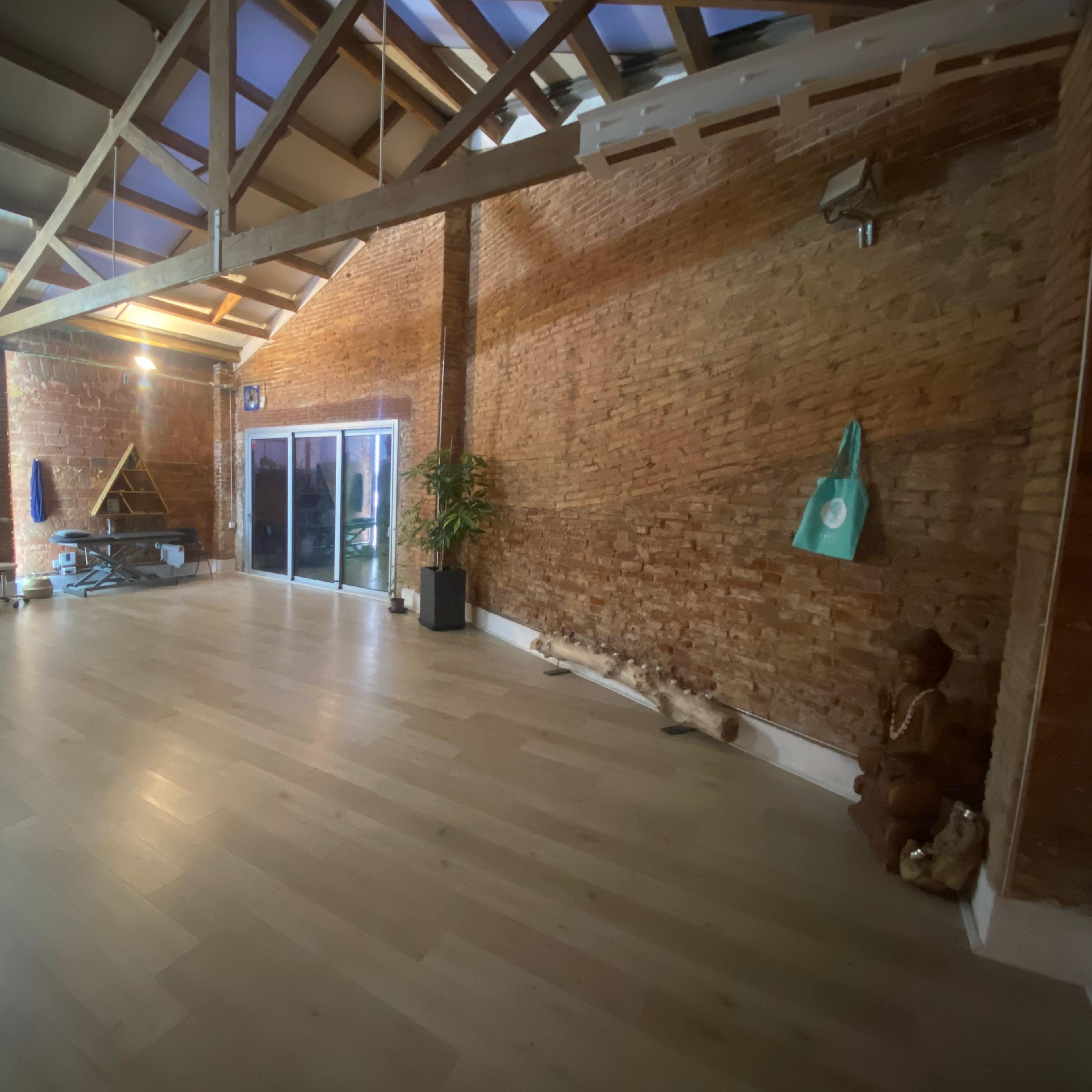 A spacious interior with exposed brick walls, wooden beams, and large glass doors leading outside, along with minimal furnishings.