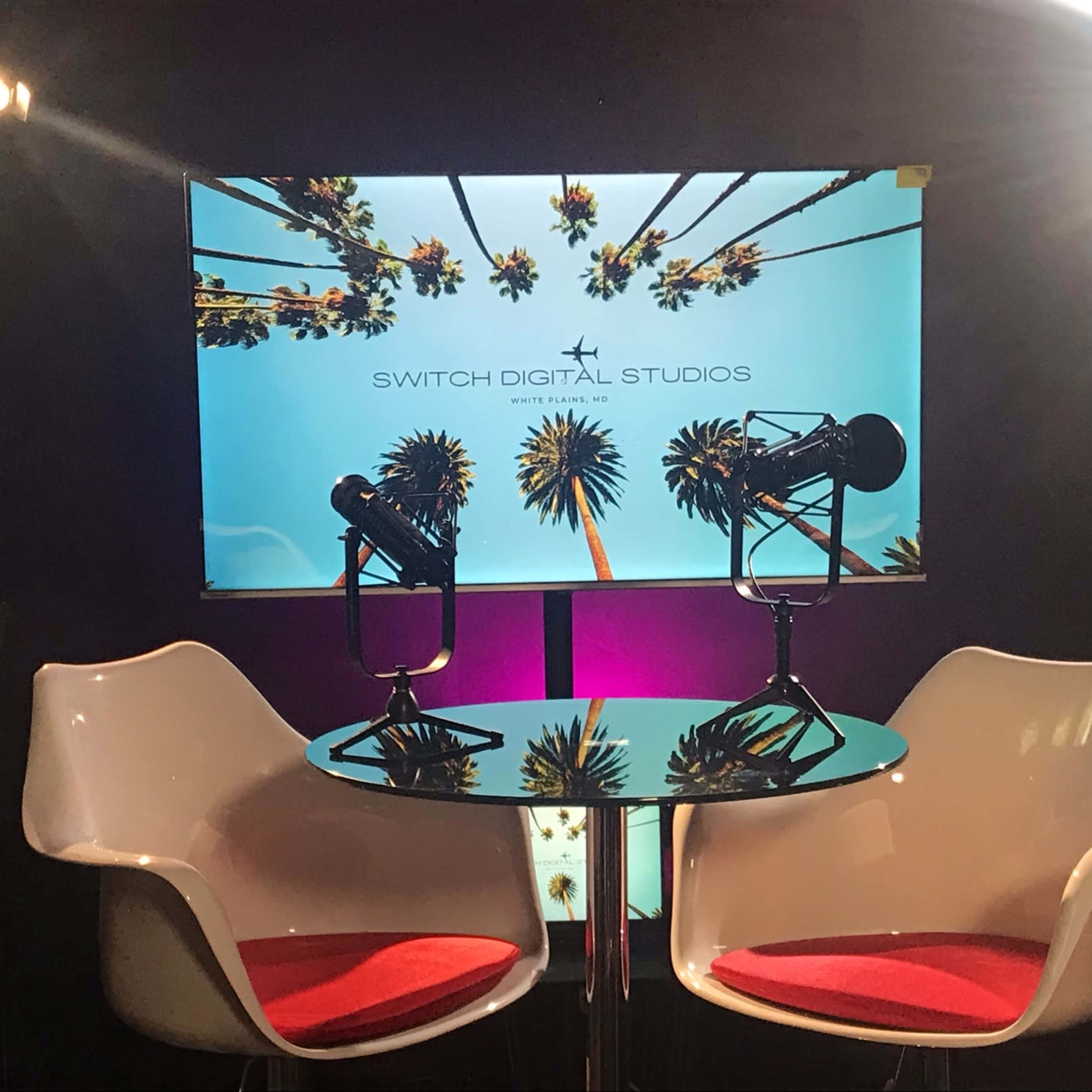 A modern studio setup featuring two white chairs, a circular table, and a backdrop with palm trees and the text "SWITCH DIGITAL STUDIOS."