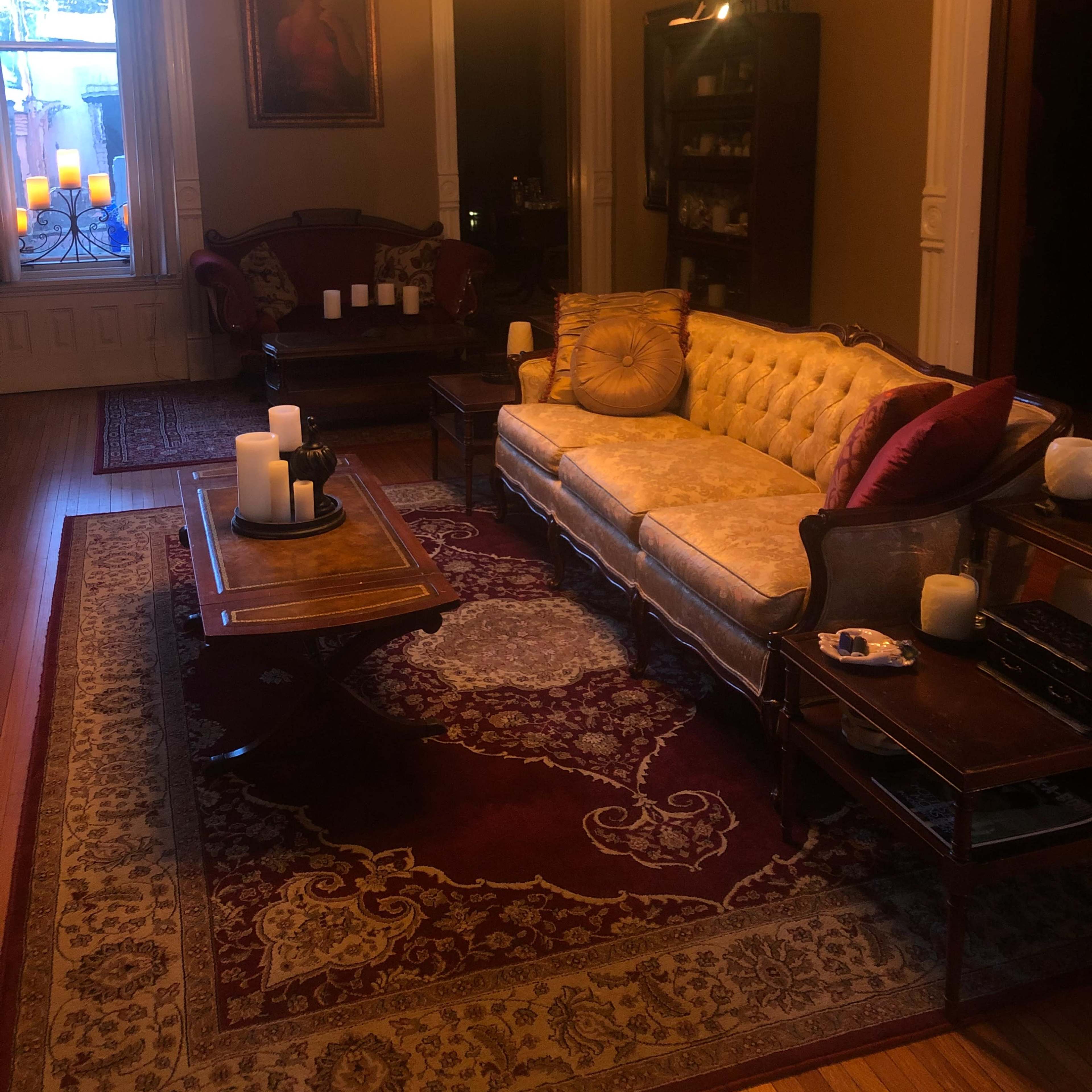 A dimly lit living room with a vintage couch, a wooden coffee table, and multiple candles arranged on furniture, set against a backdrop of ornate decor.