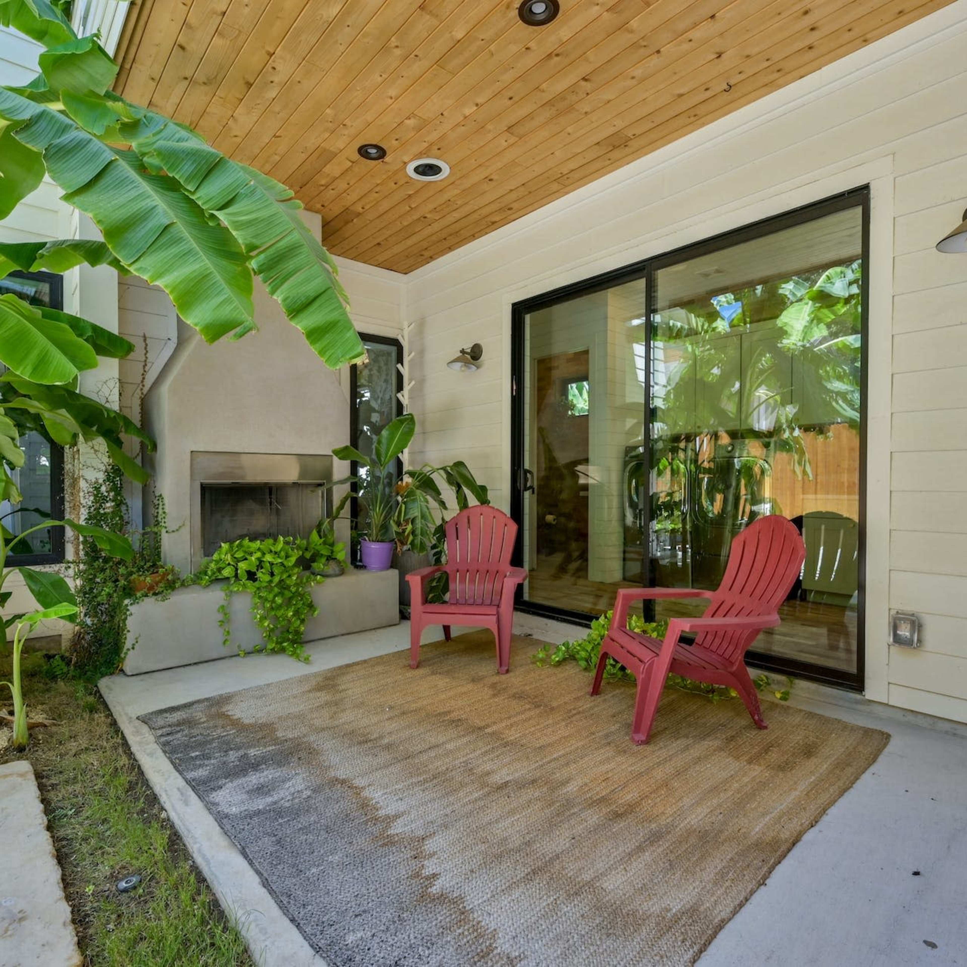 A patio area features two red chairs beside a fireplace, surrounded by lush greenery and a wooden ceiling.