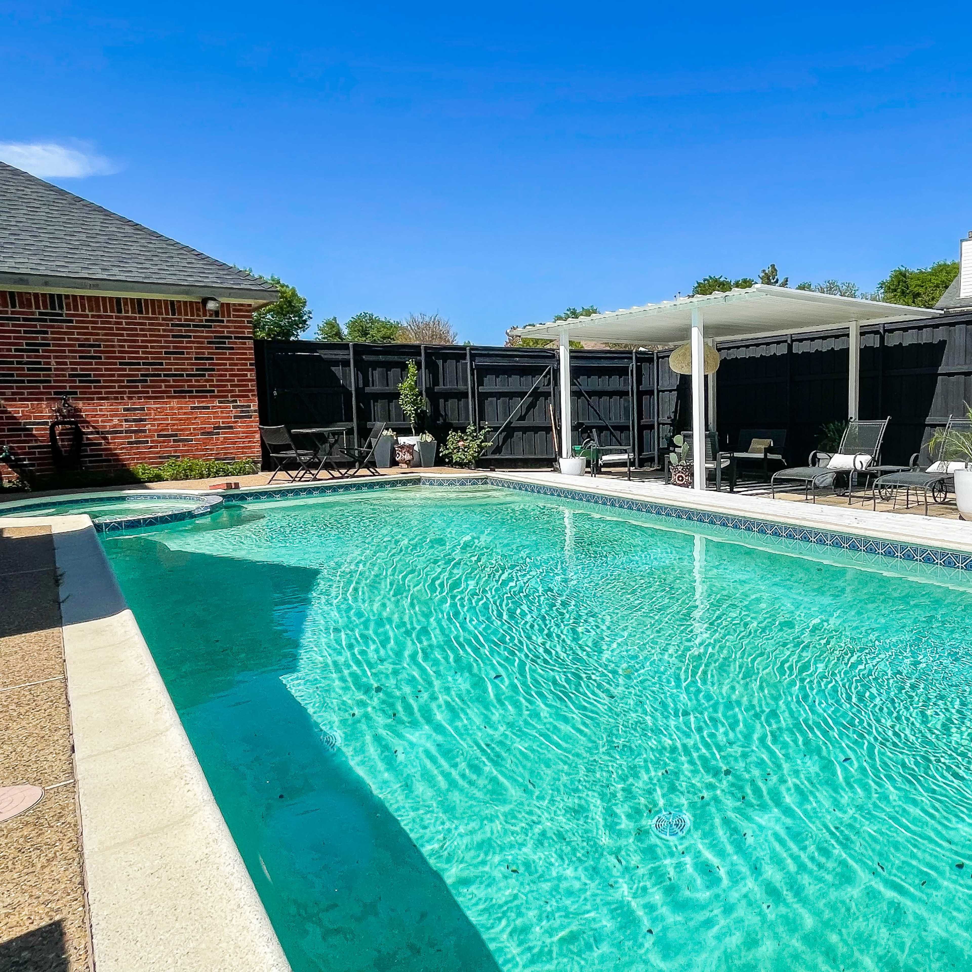 A backyard swimming pool surrounded by lounge chairs and a covered patio area, enclosed by a black fence.