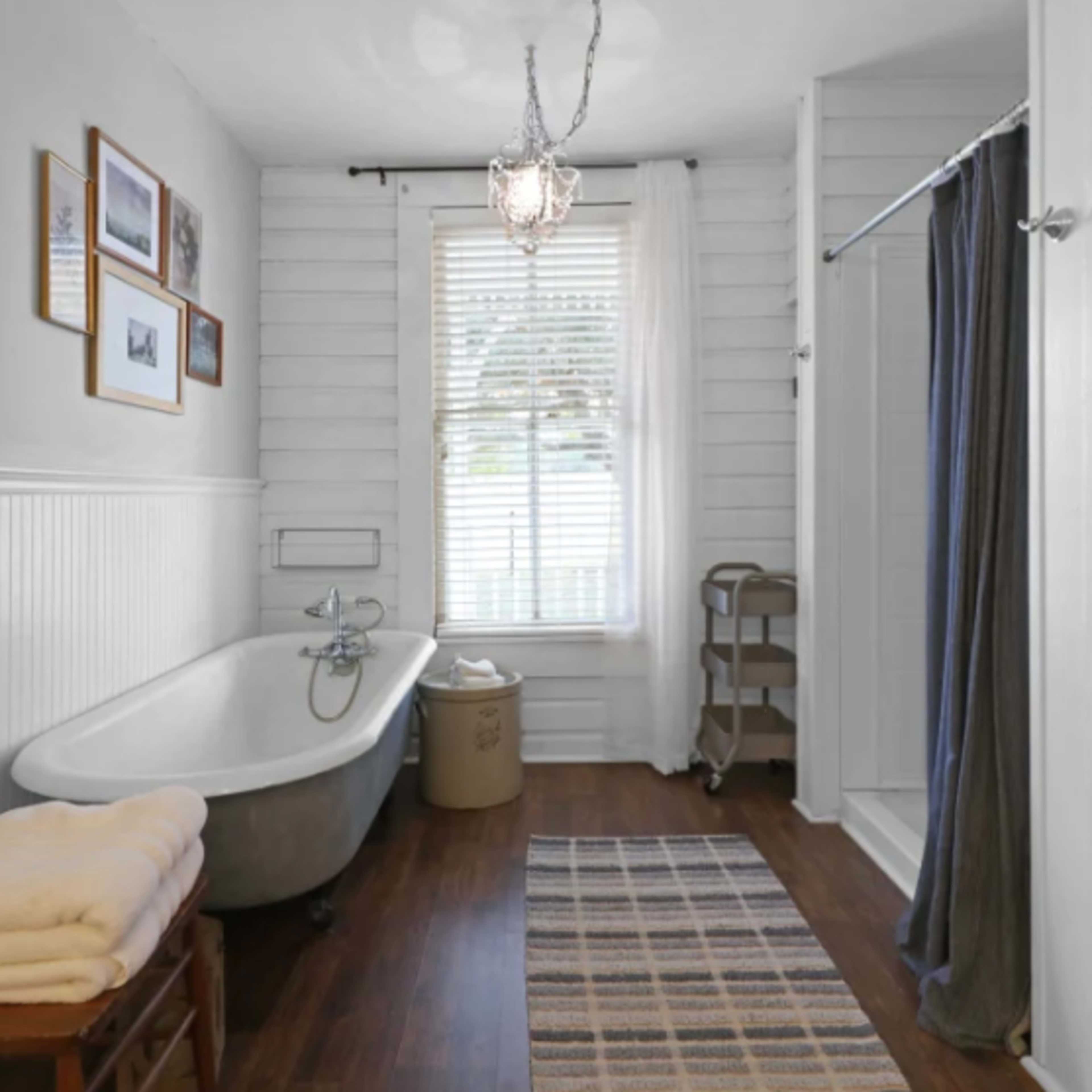 A bathroom features a freestanding bathtub, a separate shower area, a wooden chair, and a window with blinds.