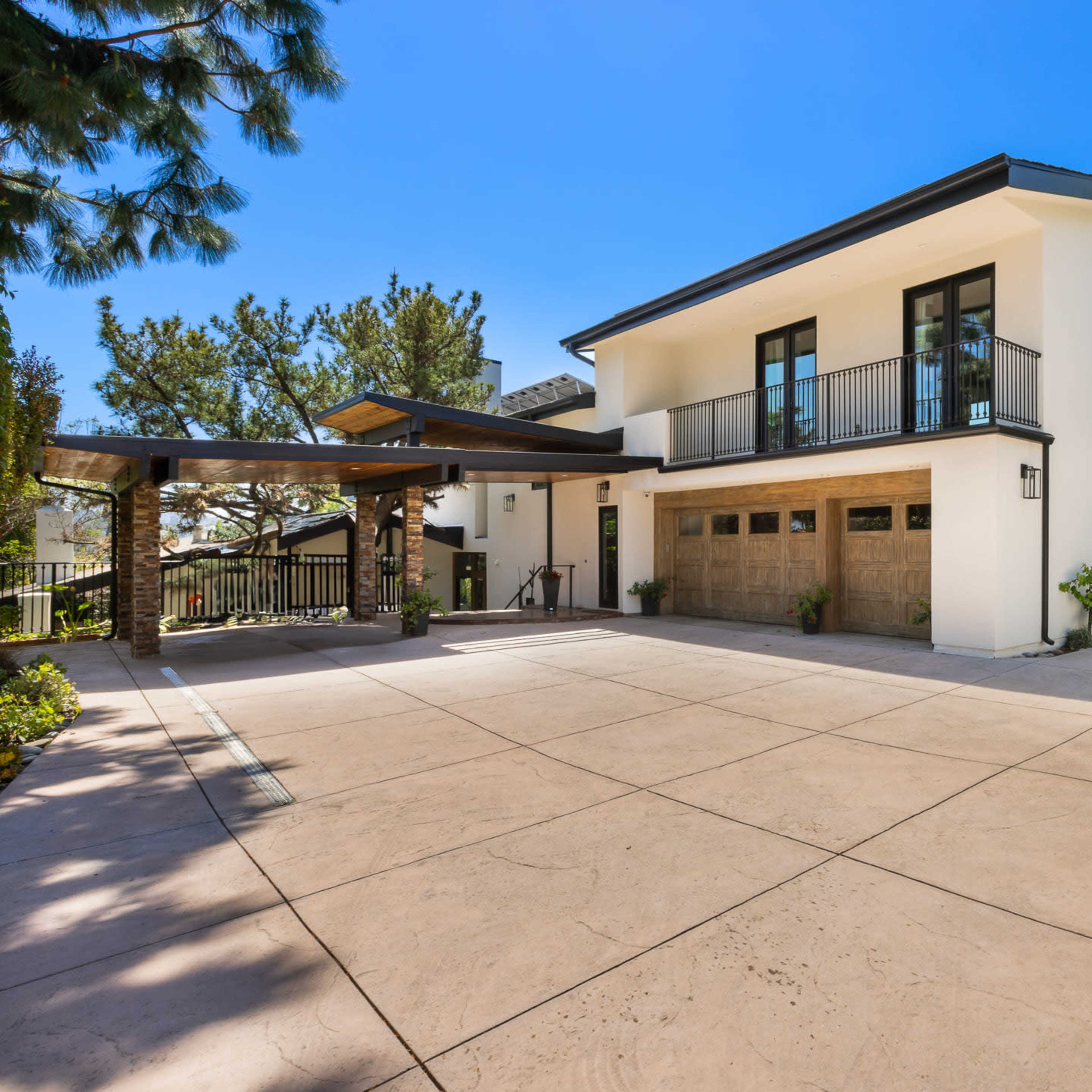 A modern two-story house with a three-car garage features a large concrete driveway and is surrounded by greenery, including a trampoline and a basketball hoop in the yard.