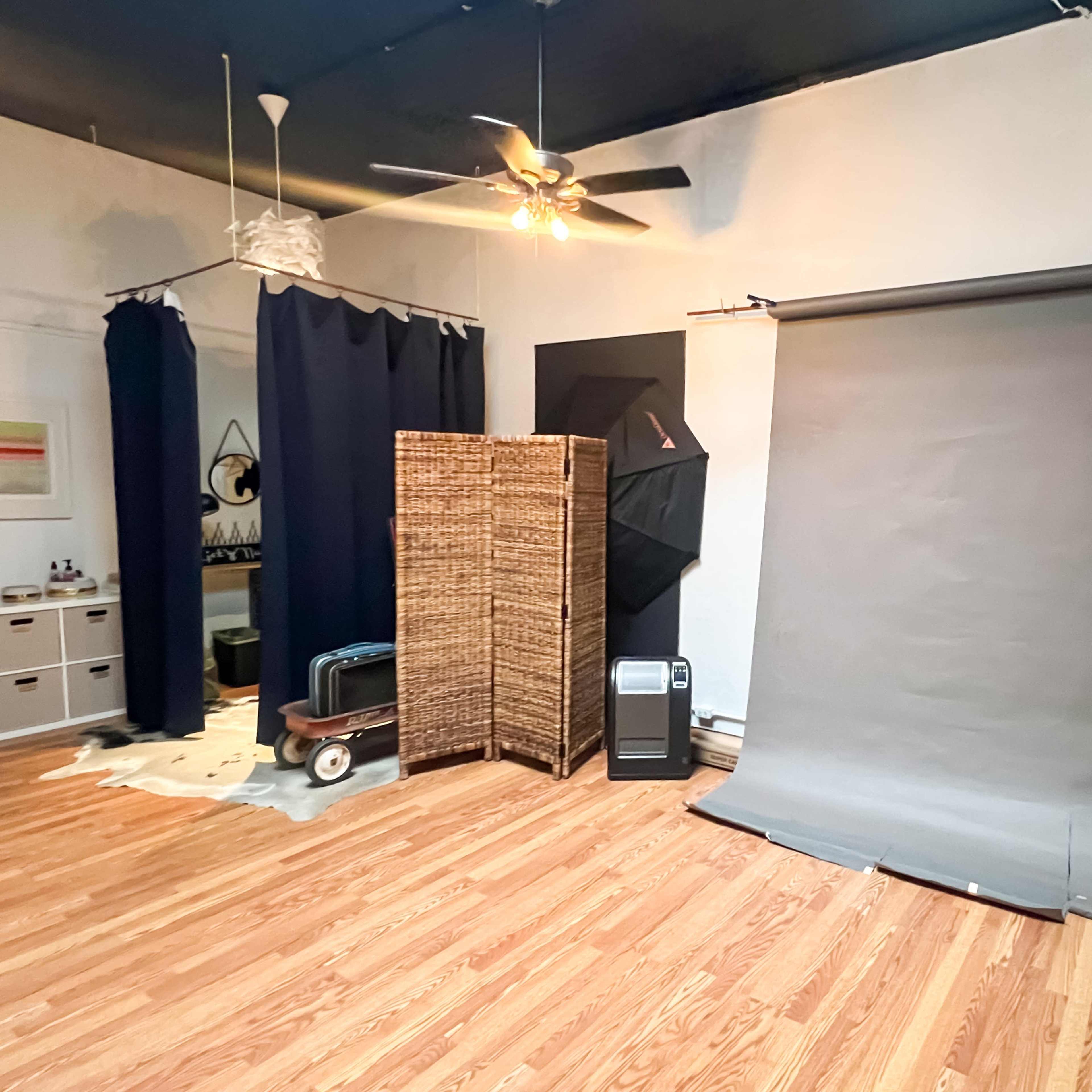 A spacious room with wooden flooring, featuring a gray backdrop, a screen divider, a fan, and various pieces of furniture.