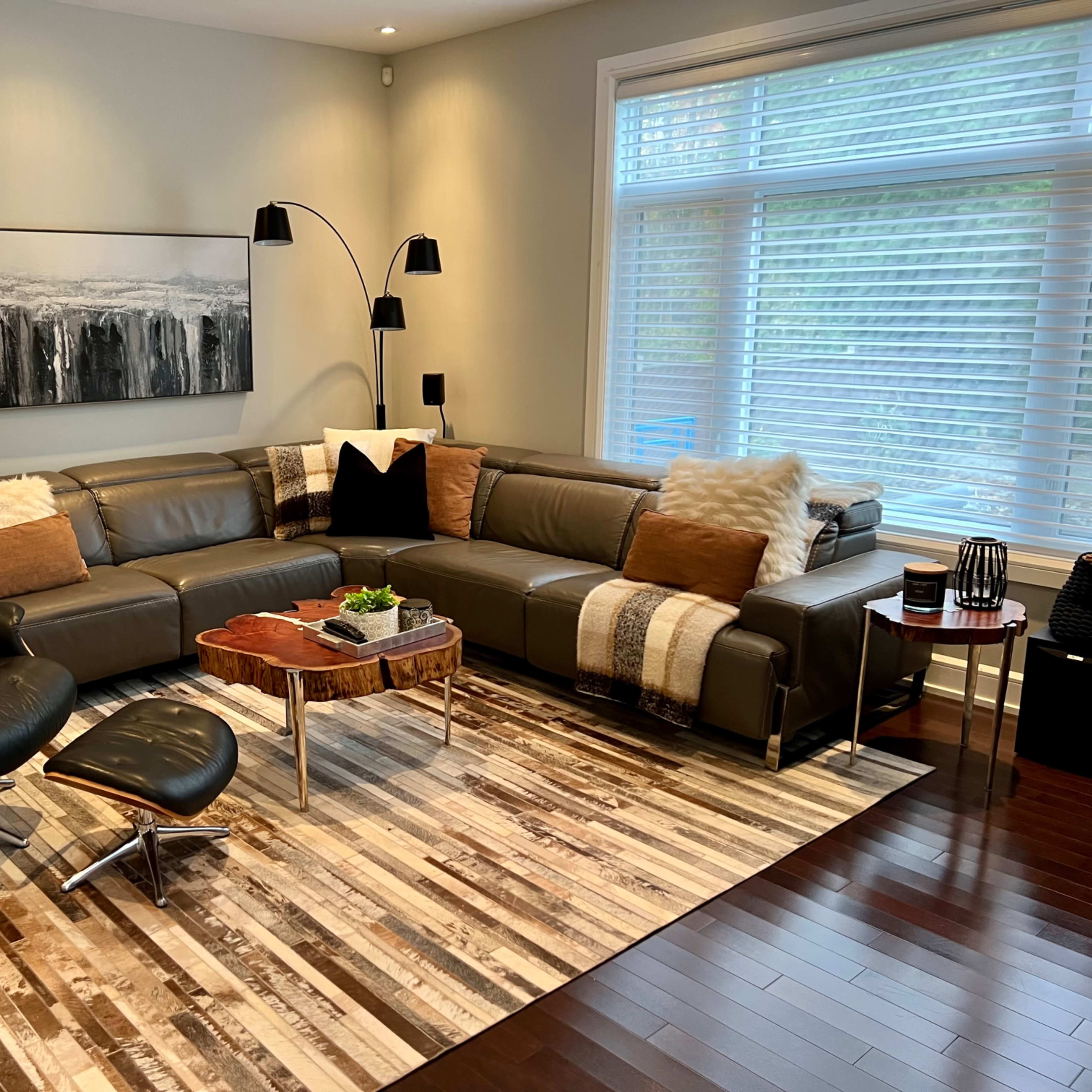 A modern living room with a large sectional sofa, a coffee table, and a lounge chair, all arranged on a patterned area rug beneath large windows with horizontal blinds.