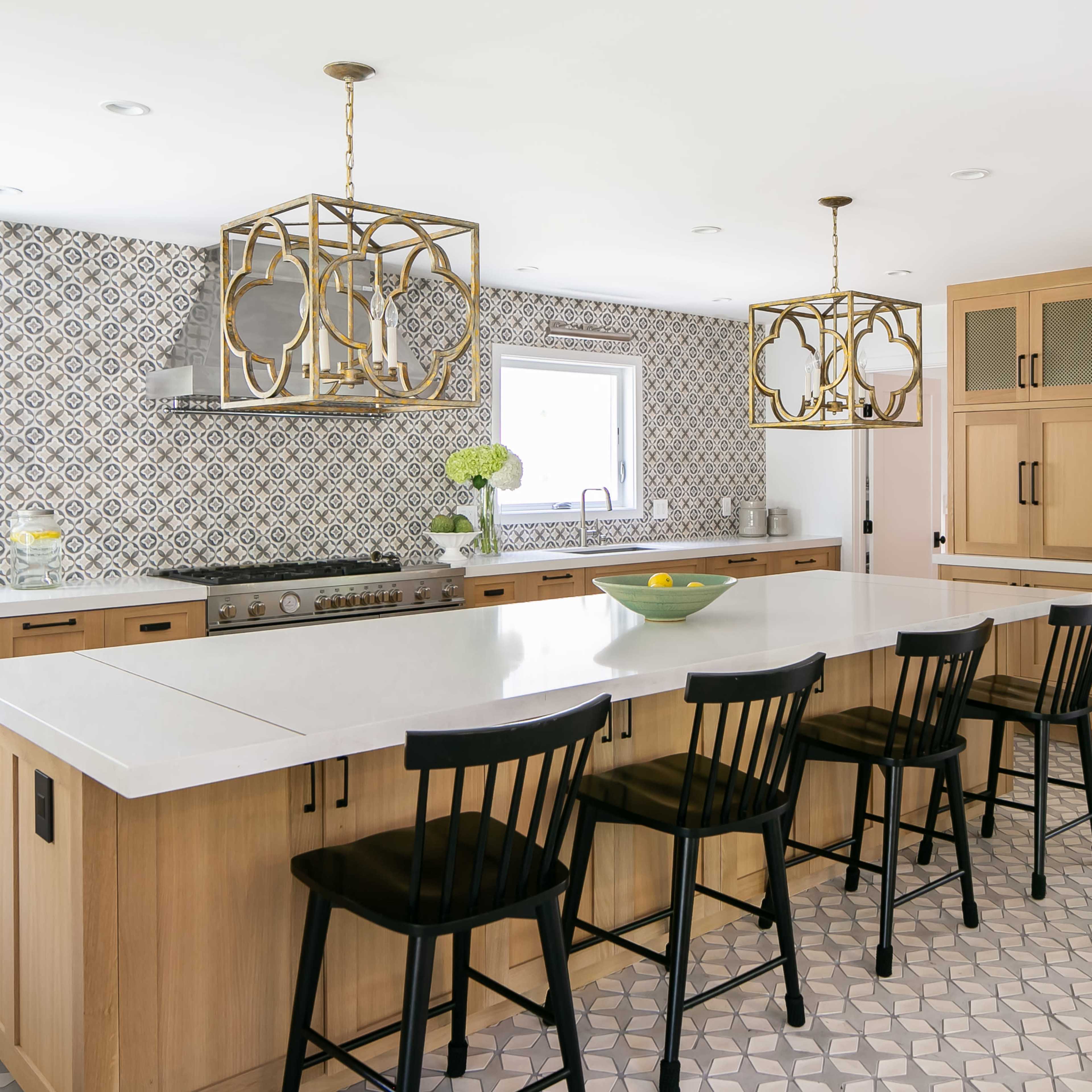 The kitchen features light wood cabinetry, a large island with seating, and decorative tile backsplashes paired with gold light fixtures.