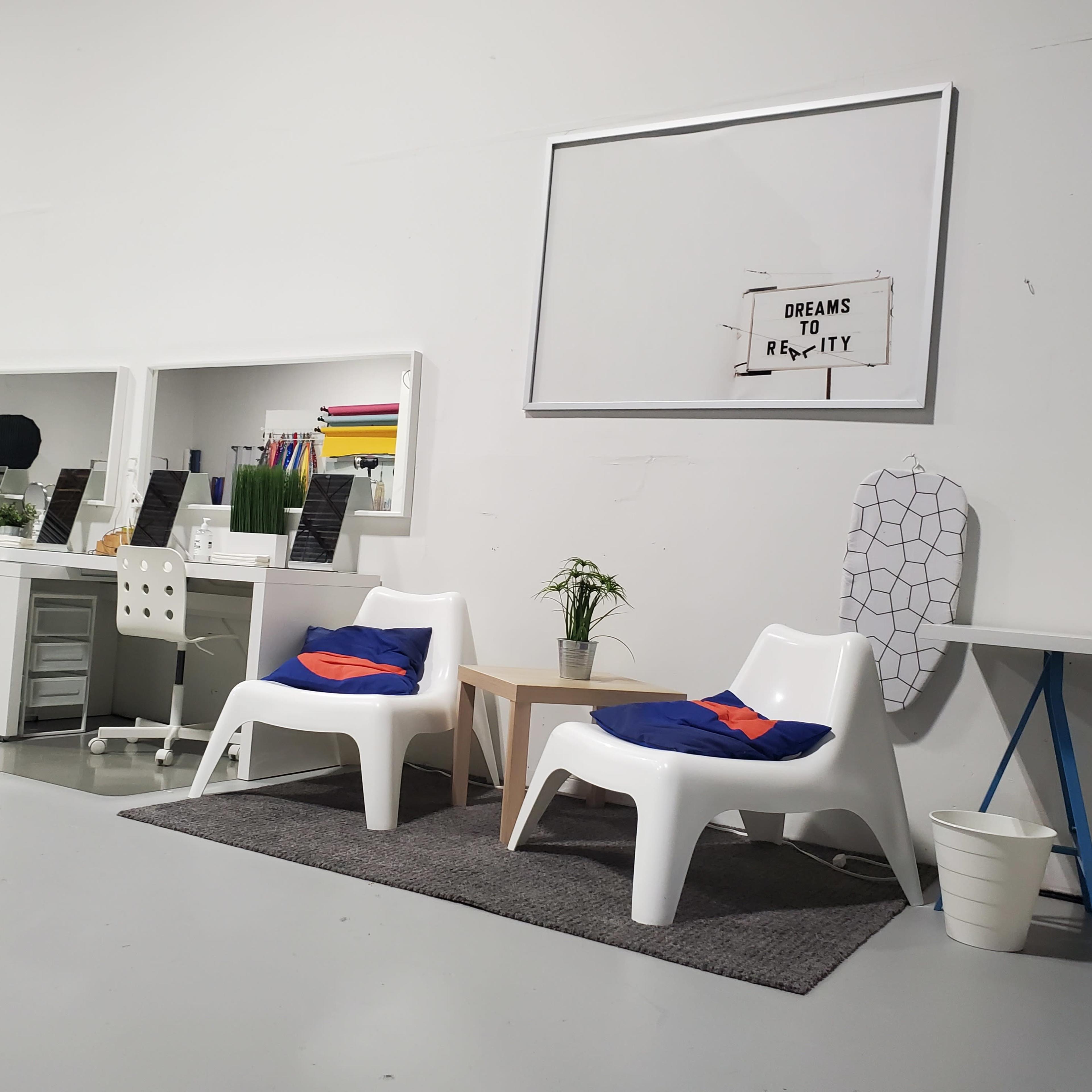 A minimalist office space featuring two white chairs, a small wooden table, potted plants, and a large empty frame on the wall.