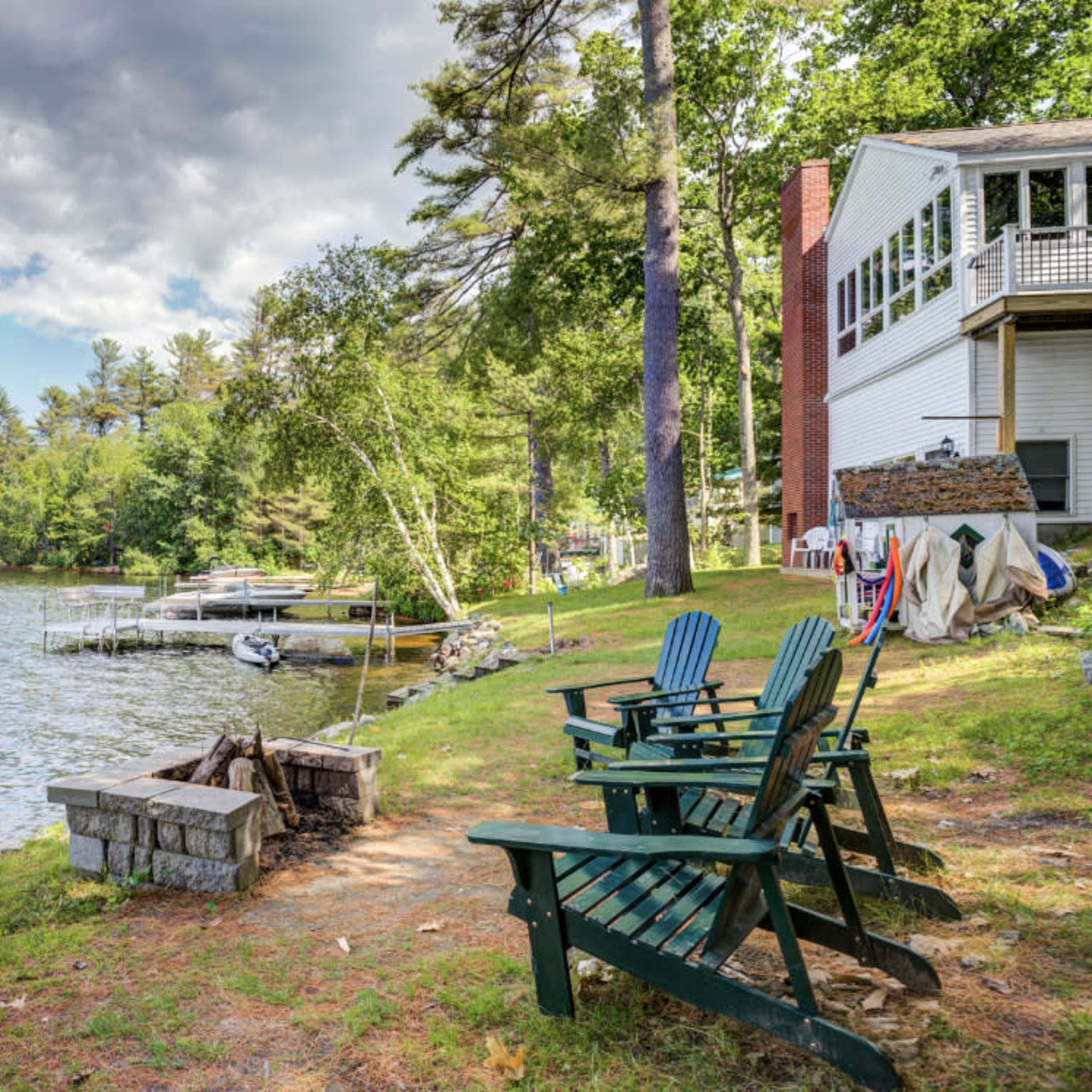 A view of a lakeside area featuring three green Adirondack chairs near a stone fire pit, with a house and dock visible in the background surrounded by trees.