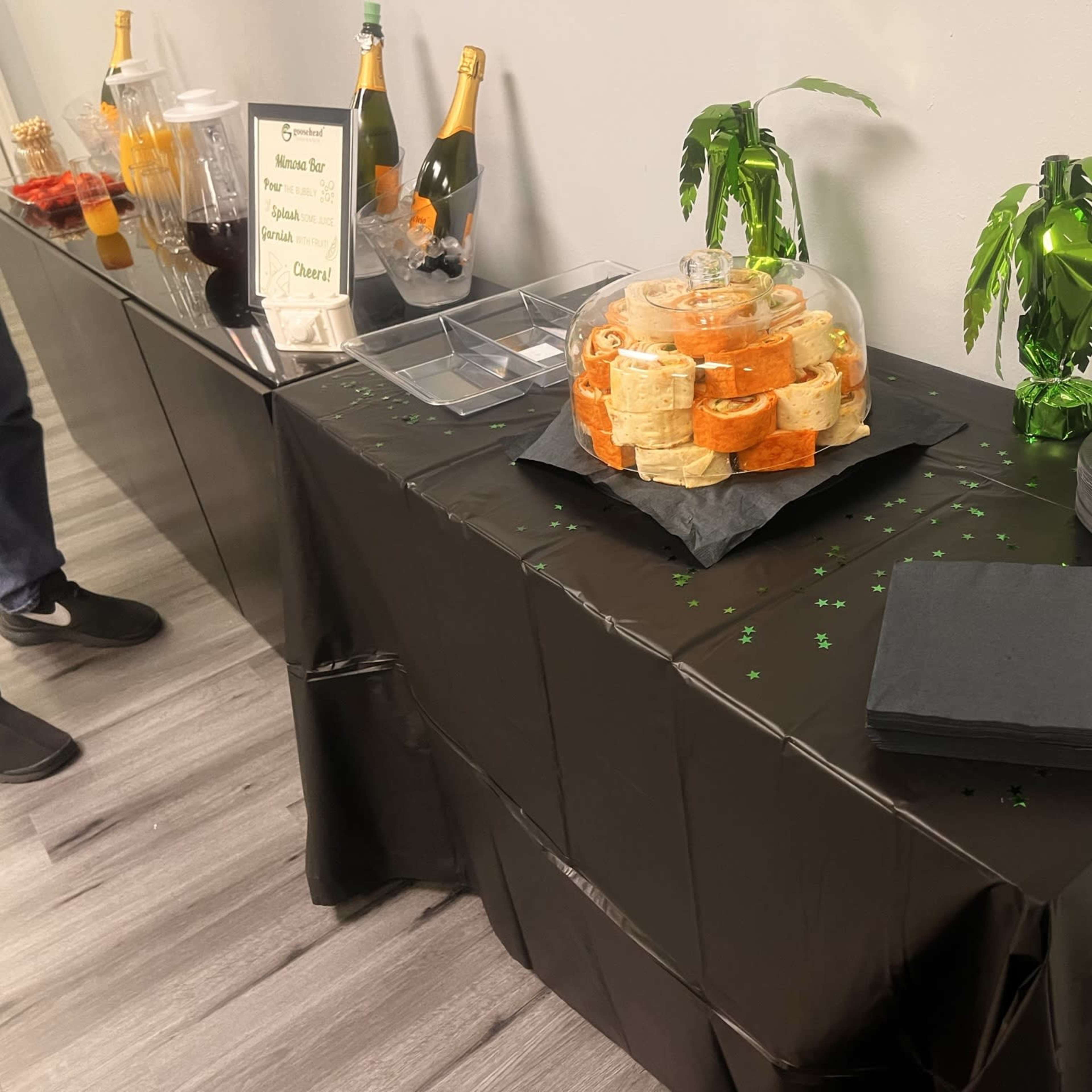 A buffet table covered with a black cloth displays a platter of assorted wraps, beverages, and decorative green palm tree centerpieces.