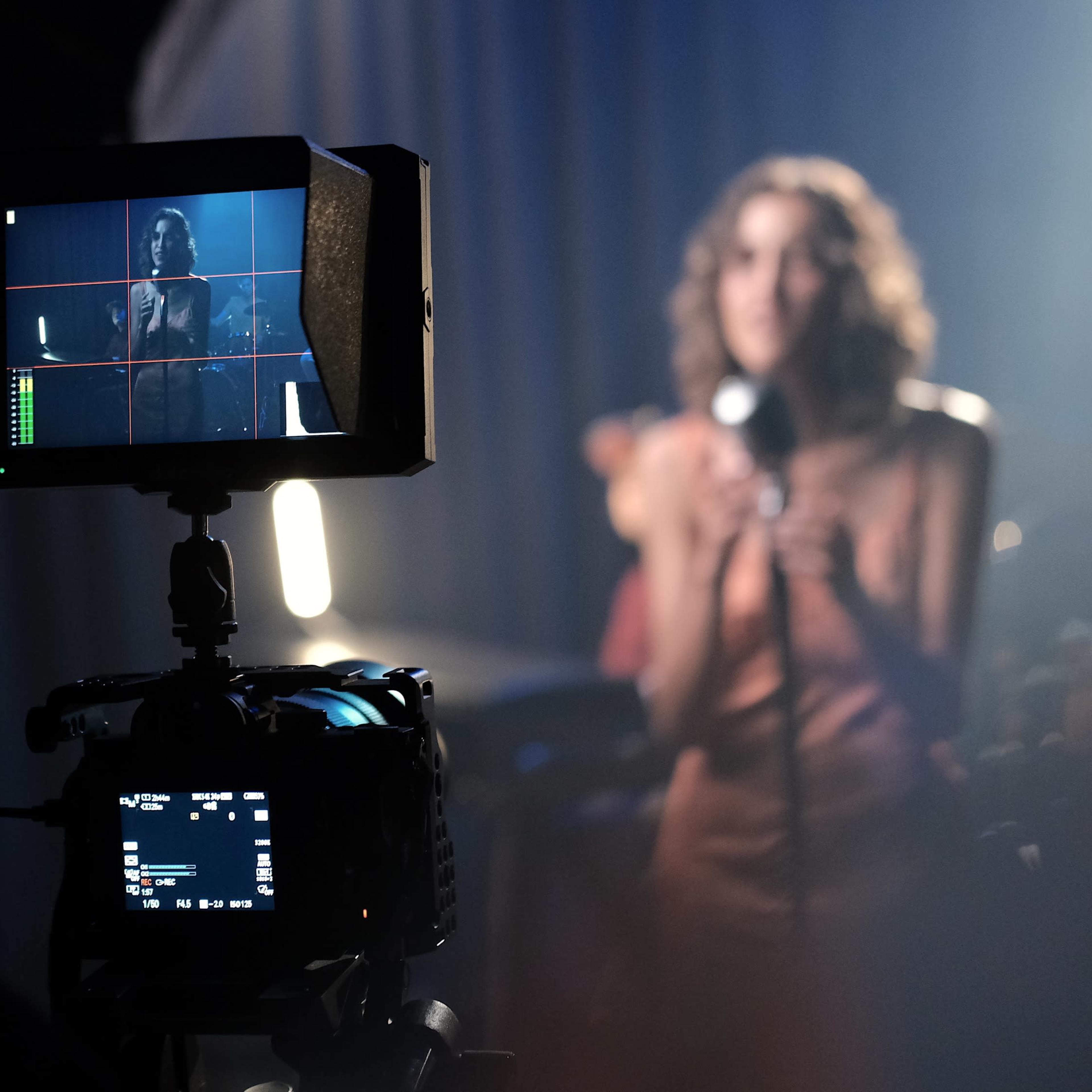 A camera operator captures a performer singing on stage, with a monitor displaying the live feed.