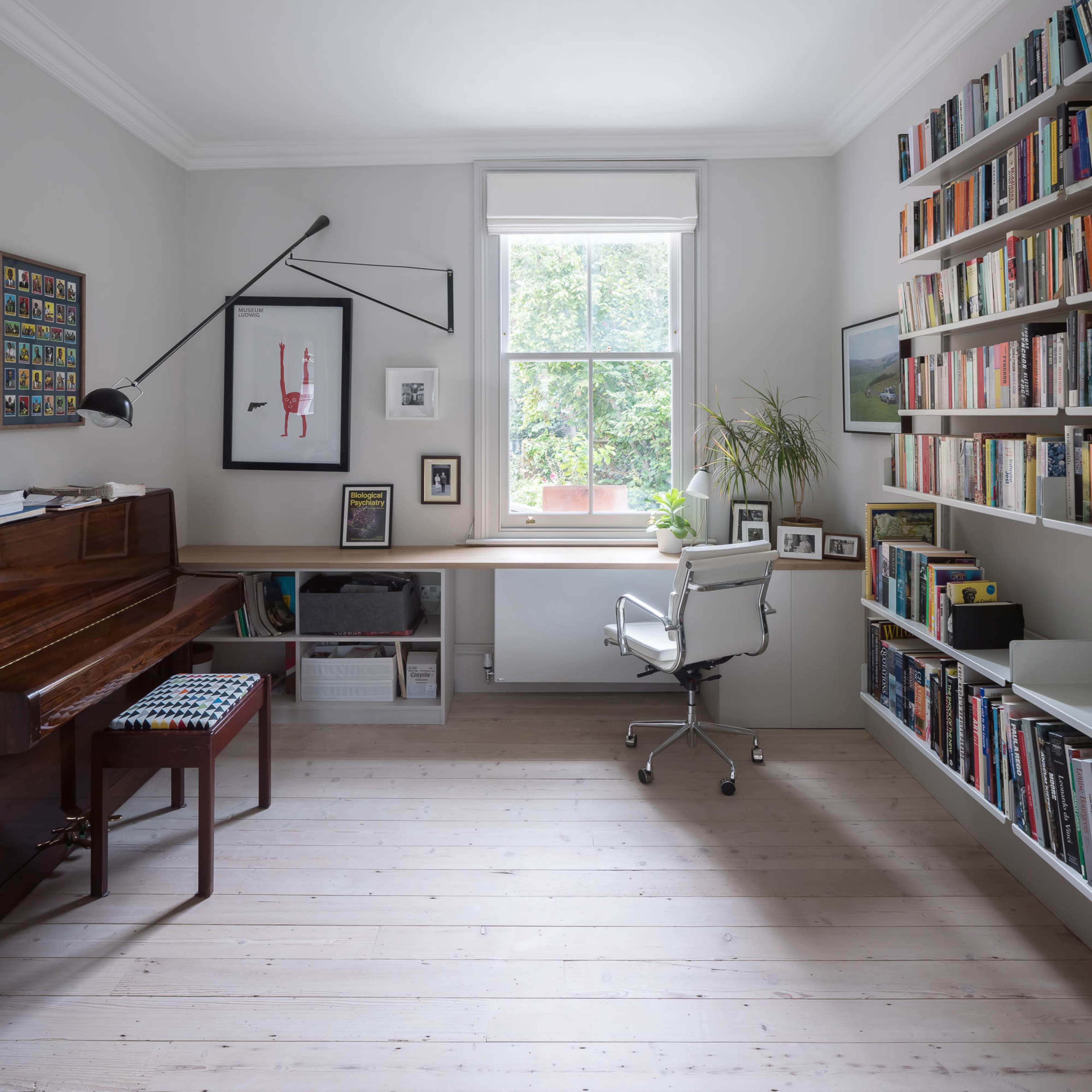 A bright home office featuring a wooden piano, a modern desk with a chair, and shelves filled with books against a light-colored wall.