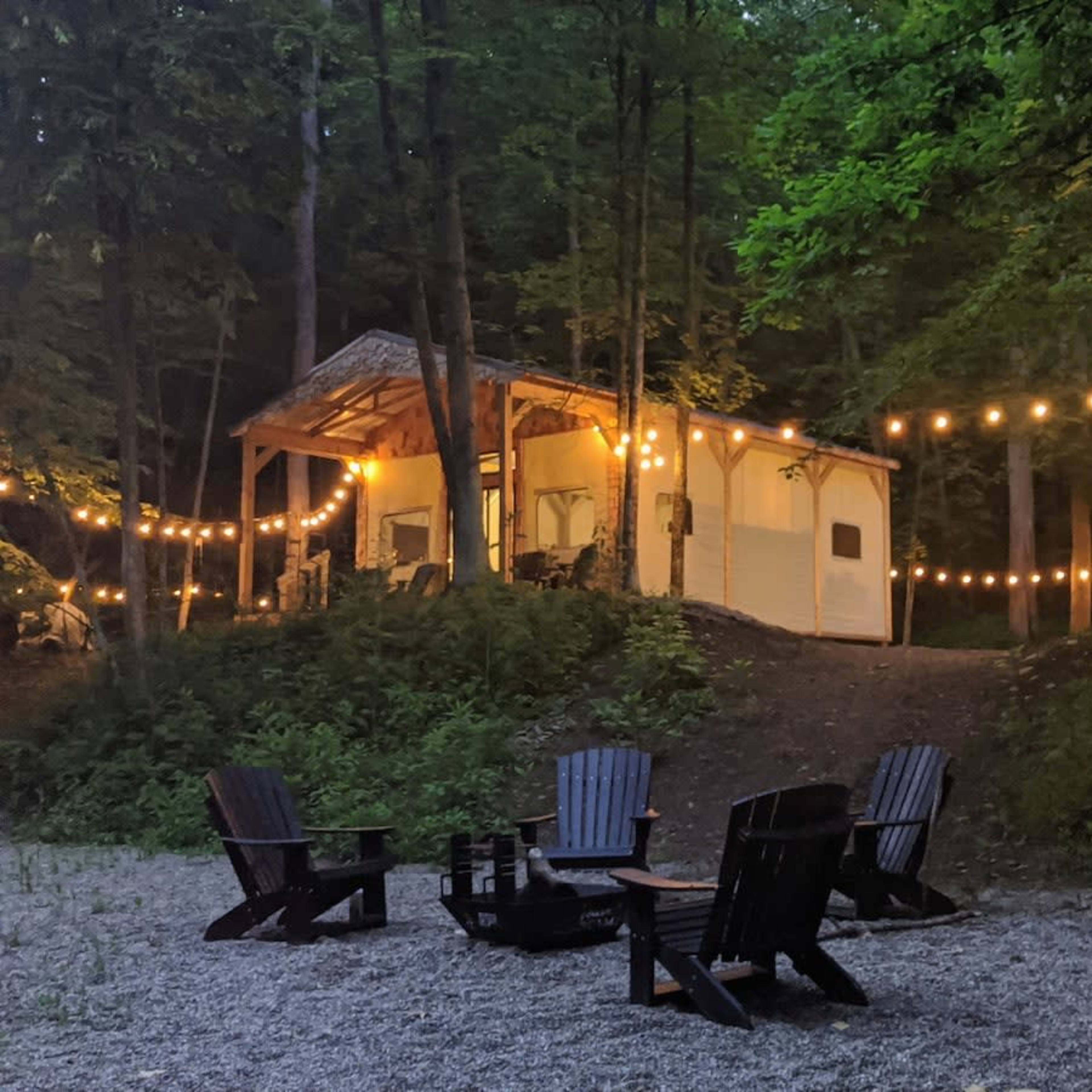 A cabin is illuminated by string lights in a wooded area, with a fire pit surrounded by adirondack chairs in the foreground.