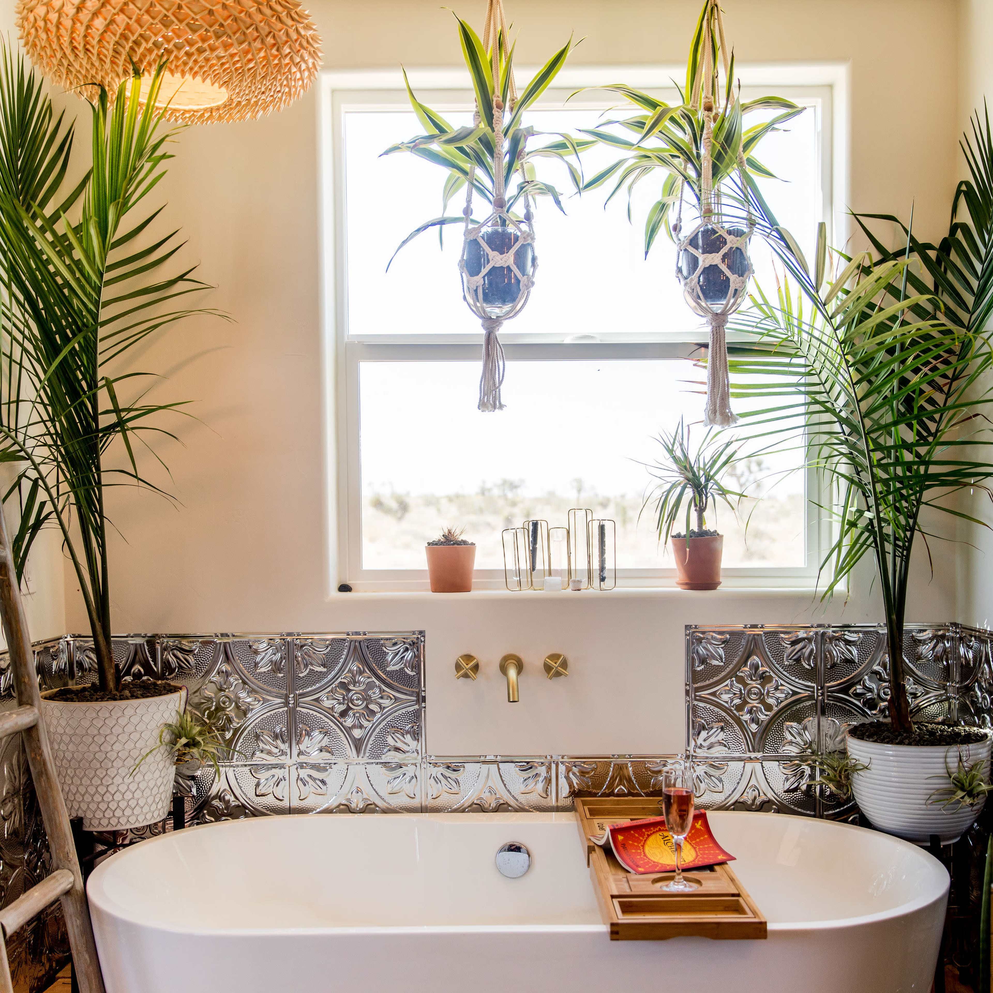 A modern bathroom features a freestanding white tub with a wooden tray, surrounded by plants and decorative hanging planters near a window.