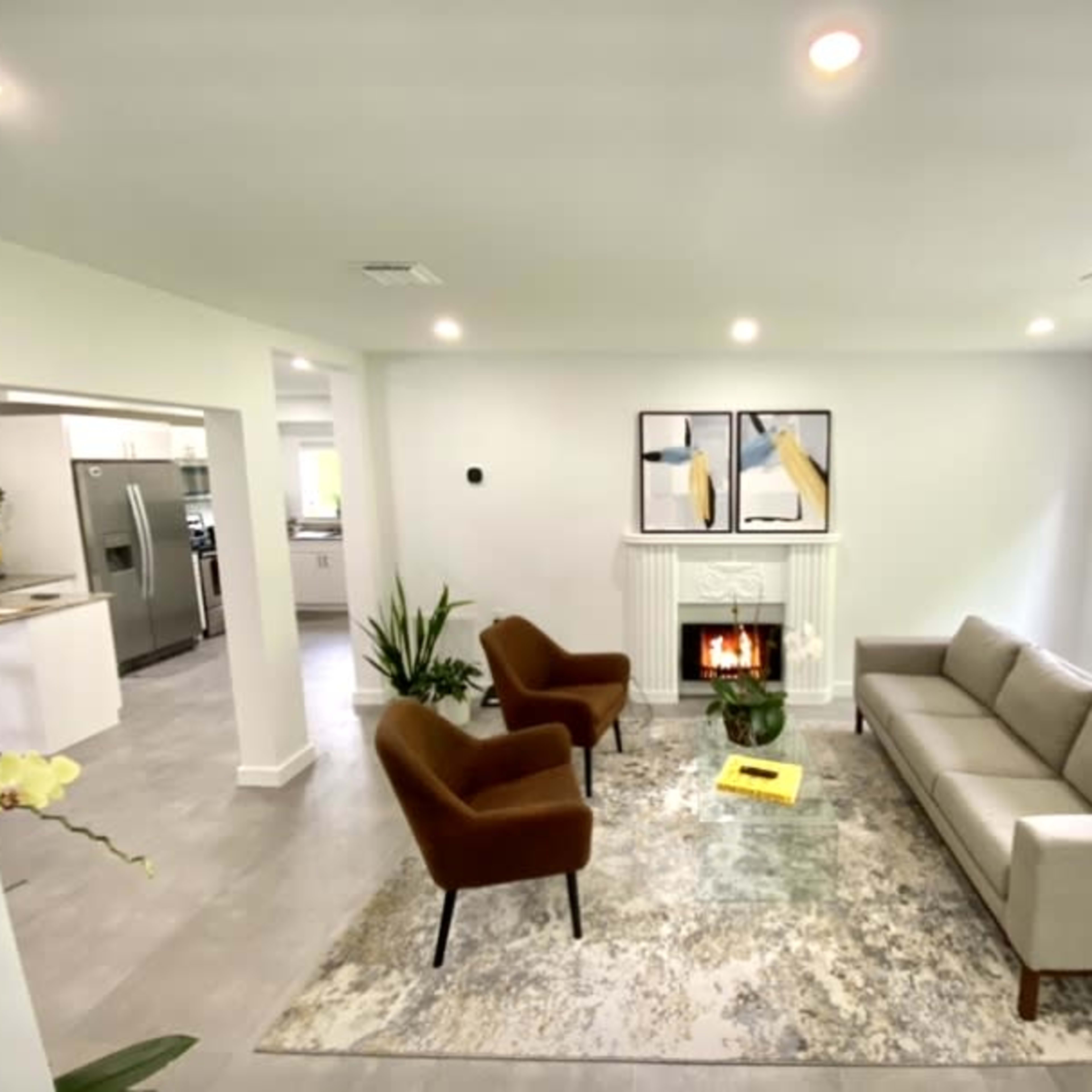 A modern living room with a sofa and two armchairs arranged around a rug near a fireplace, with large windows providing natural light.