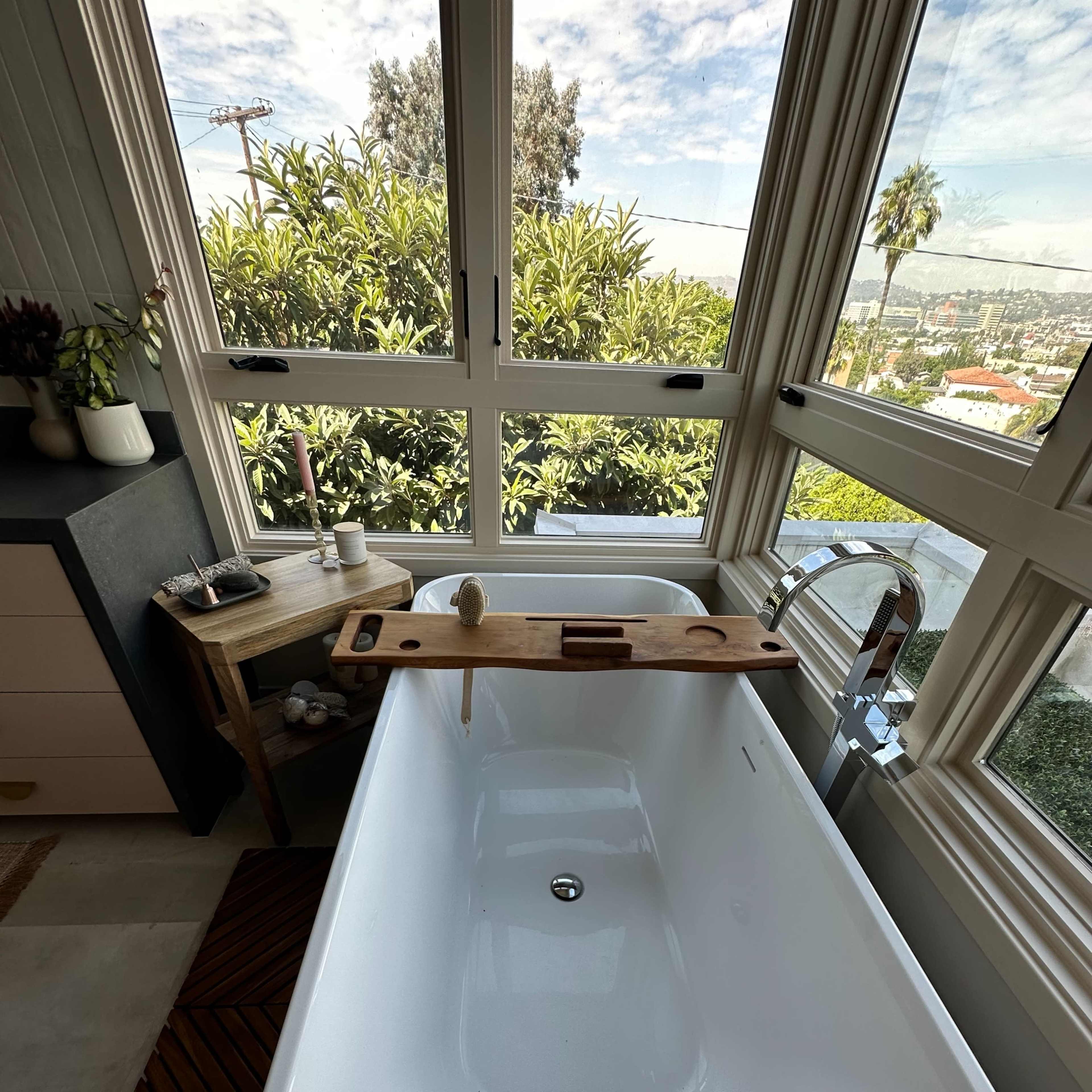 A modern bathtub with a wooden tray resting across its edge, situated in a room with large windows that provide a view of greenery outside.