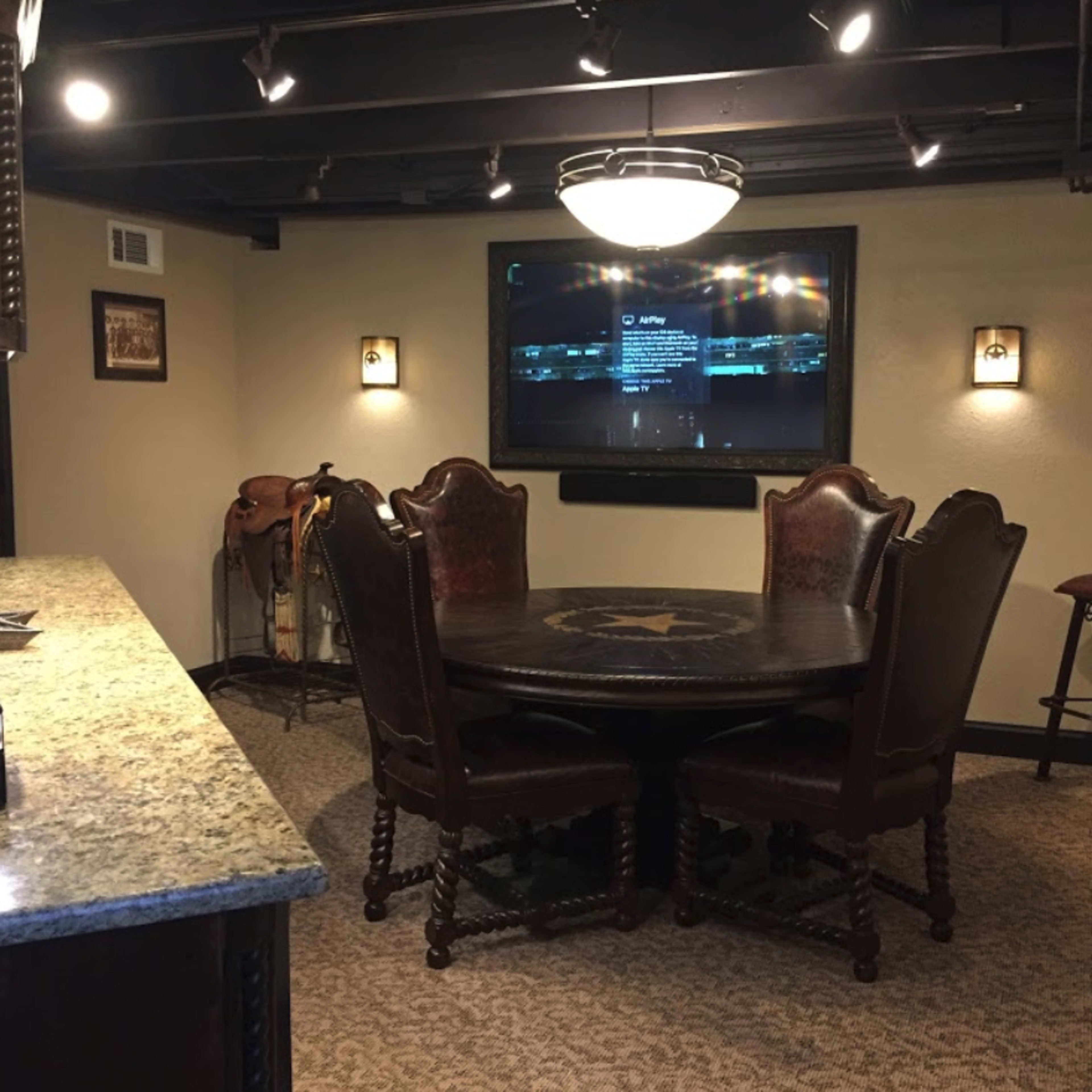 A well-lit room featuring a round table surrounded by six chairs, a mounted television on the wall, and a countertop on one side.