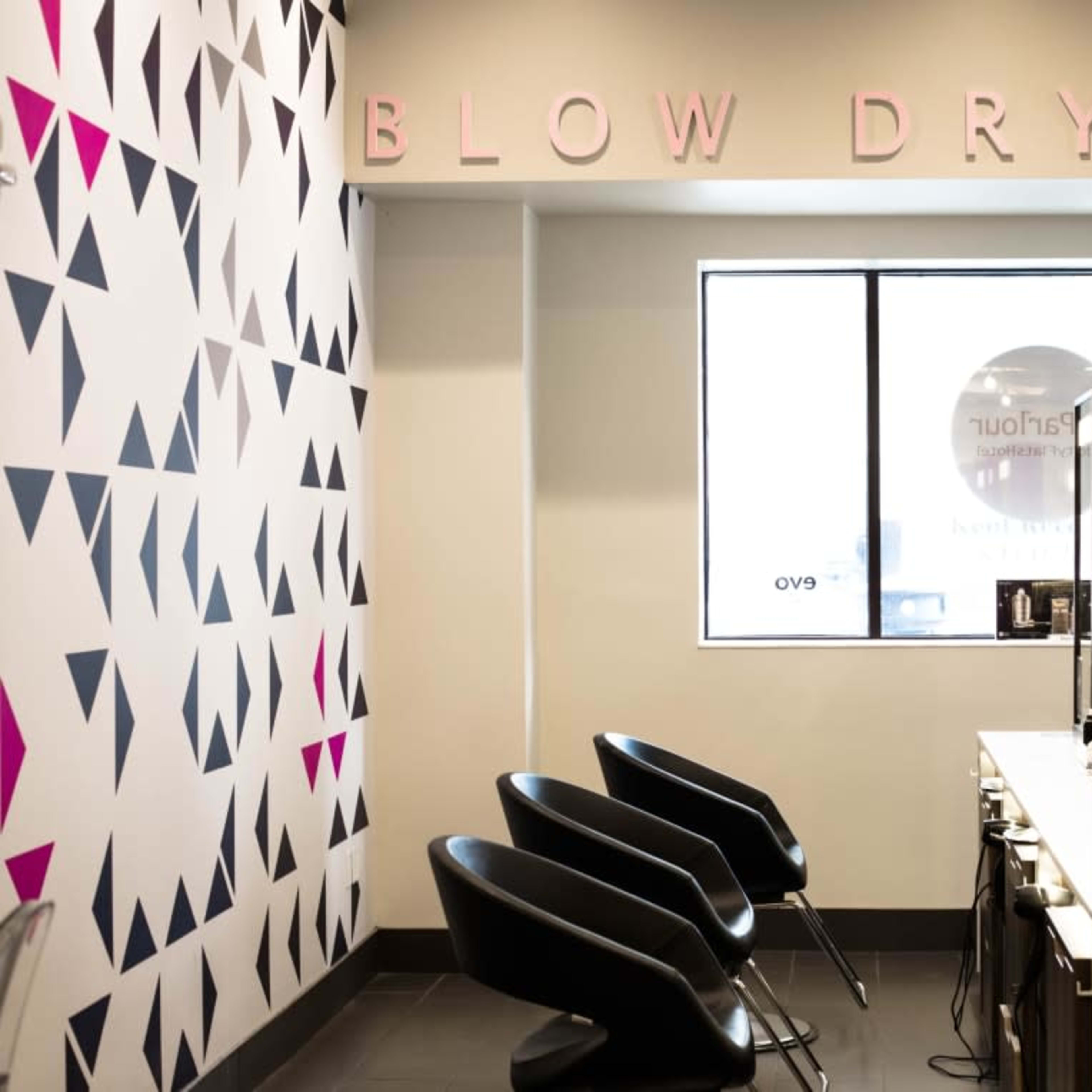 The interior of a blow dry bar featuring a decorative wall, black salon chairs, and a row of stylist stations with mirrors.