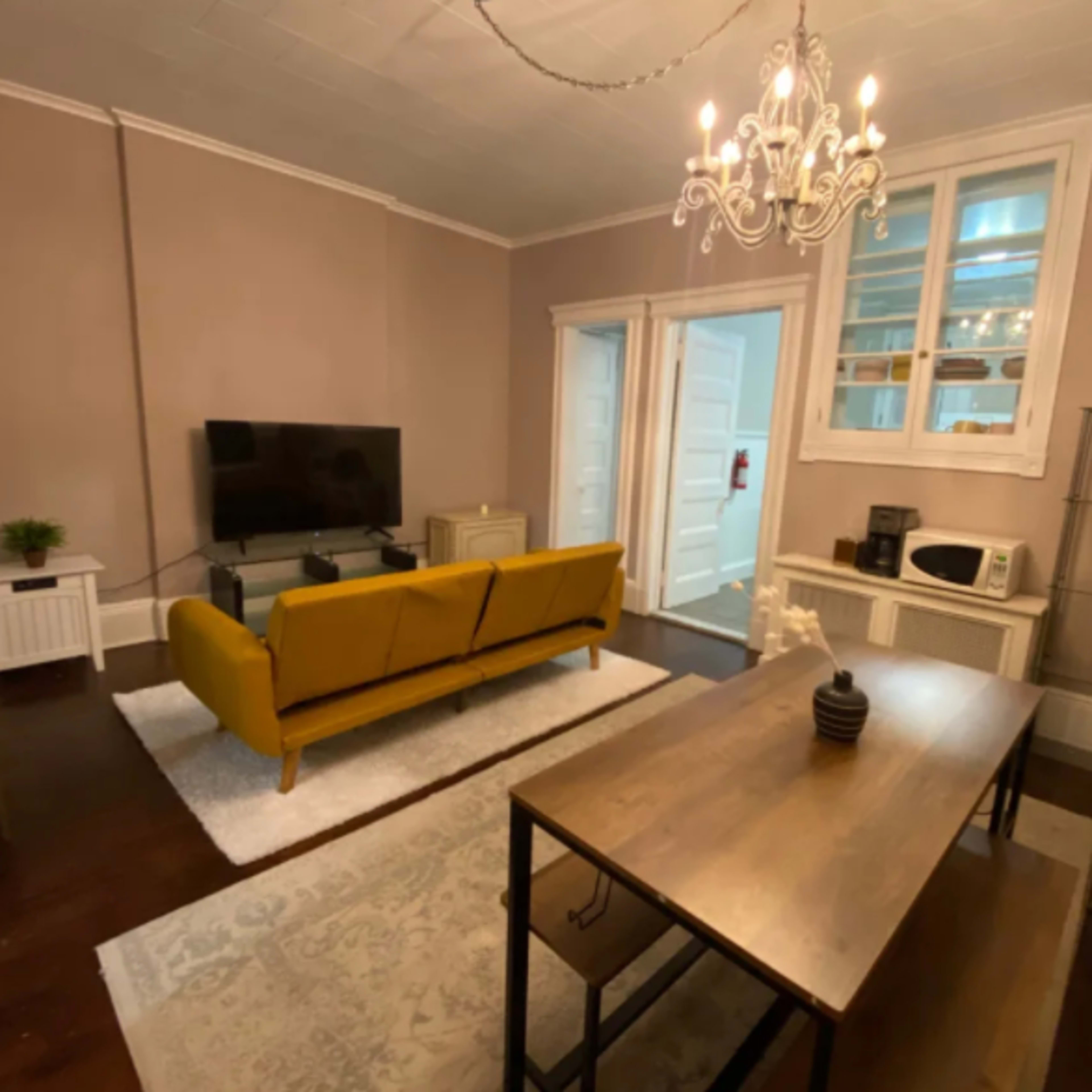 A living room with a yellow couch, a television mounted on the wall, a wooden table, and a microwave on a side table, illuminated by a chandelier.