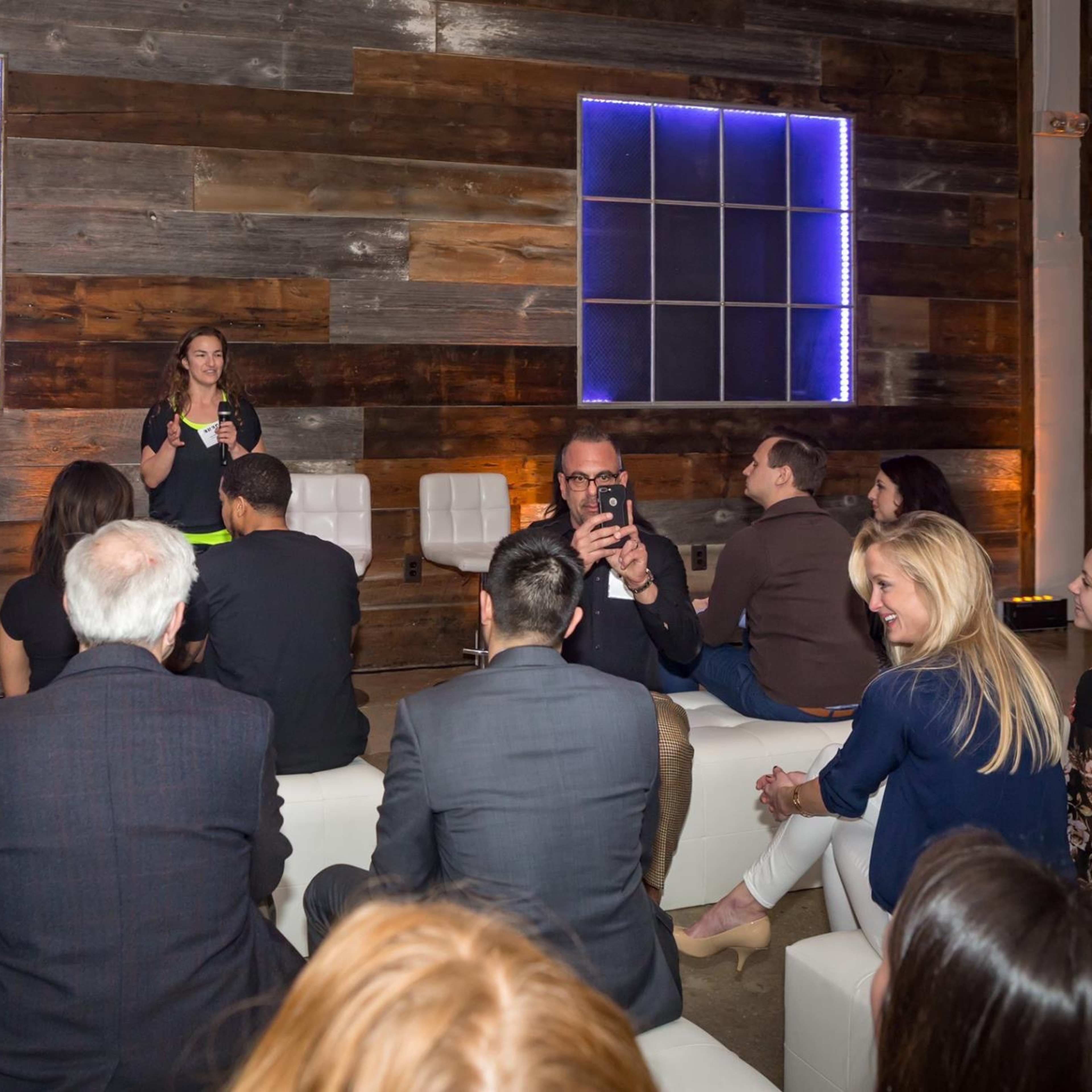 A speaker addresses an audience seated on white sofas in a stylish interior with wooden walls and glowing blue lights.