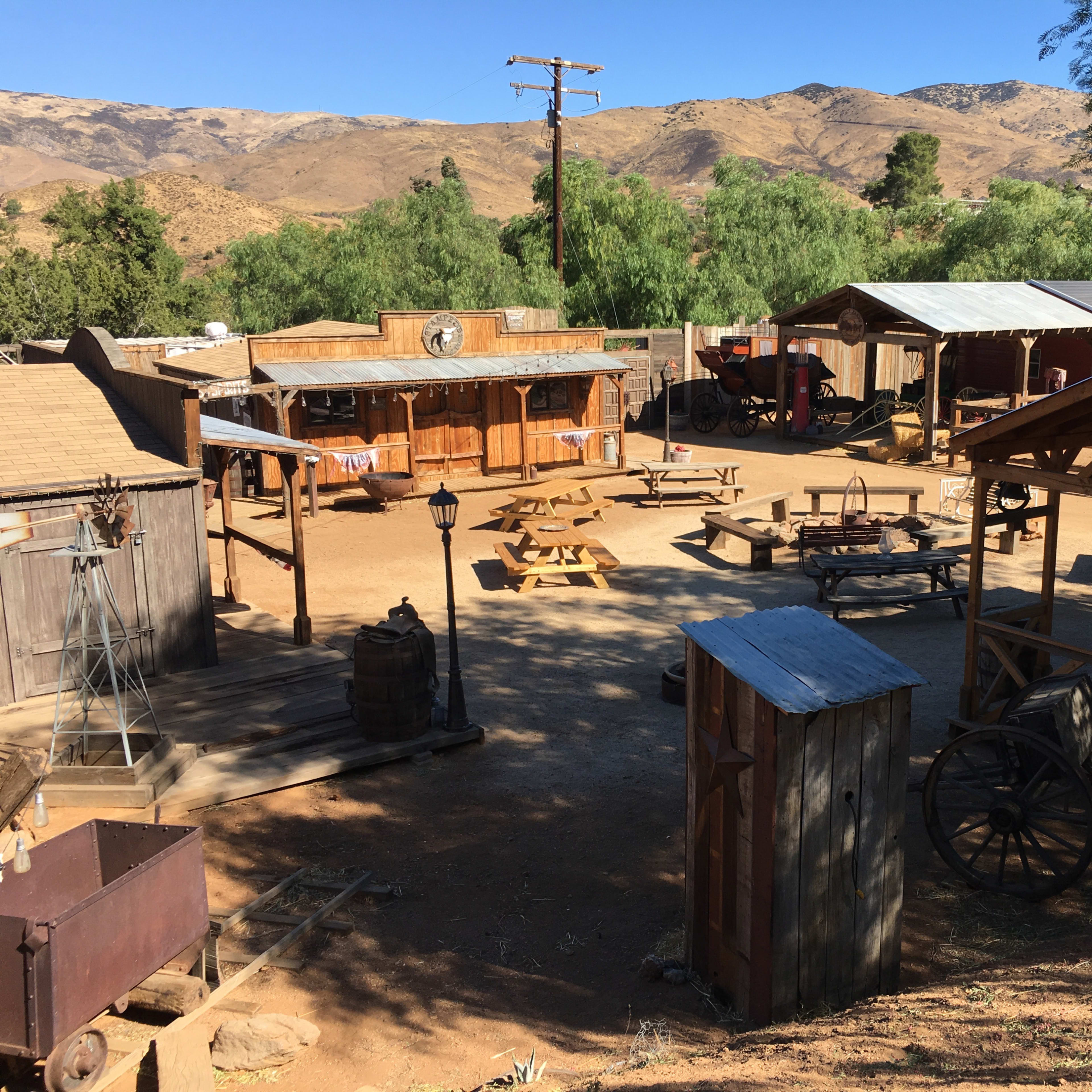 A rustic western-style movie set with wooden buildings, outdoor seating, and surrounding mountains.