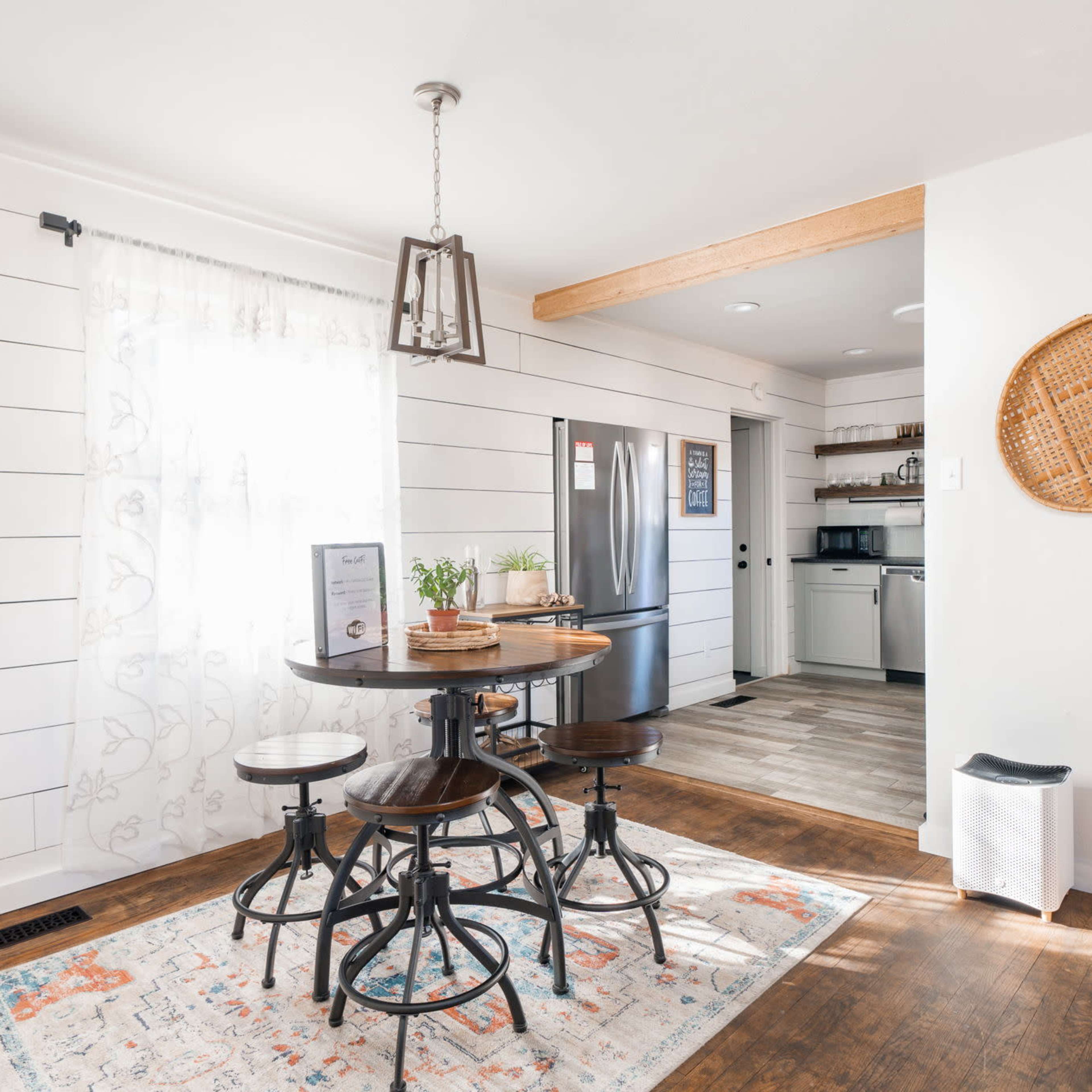 A bright kitchen with a round wooden table and four chairs, surrounded by white shiplap walls and a stainless steel refrigerator in the background.