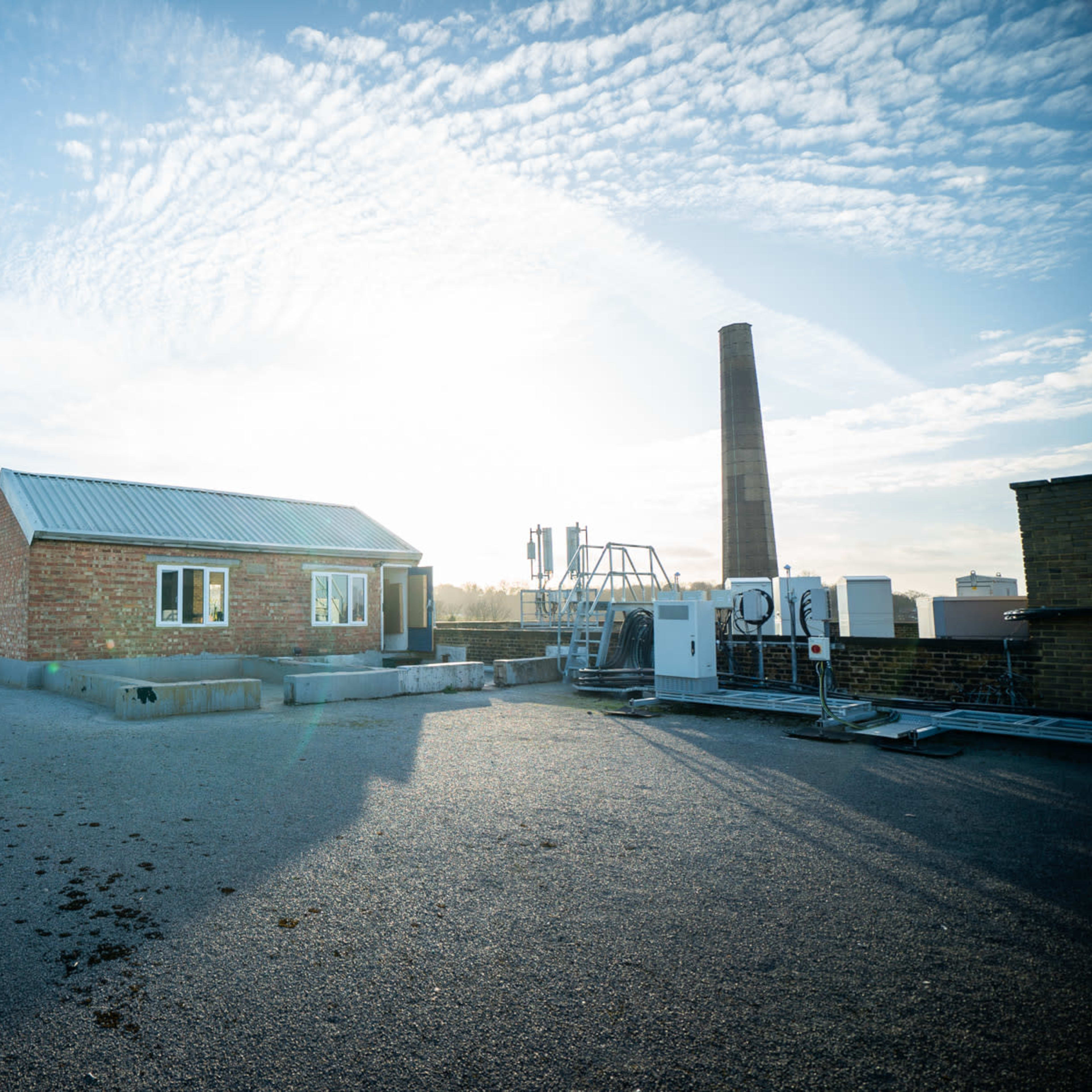 A flat rooftop with a small brick building, a chimney, and various mechanical equipment under a blue sky with scattered clouds.