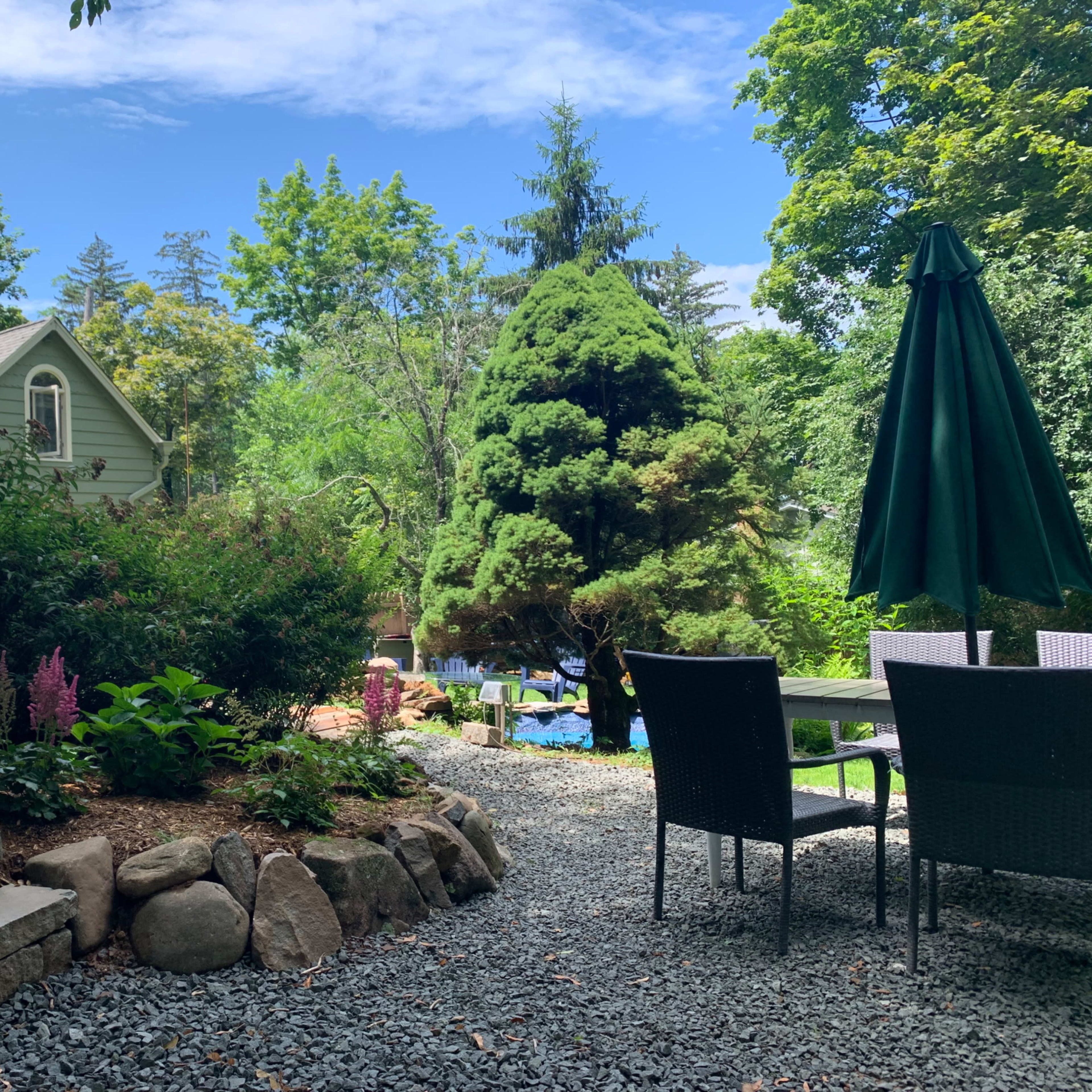 A stone path leads through a garden area with greenery and a patio set beneath a large umbrella, alongside a house in the background.