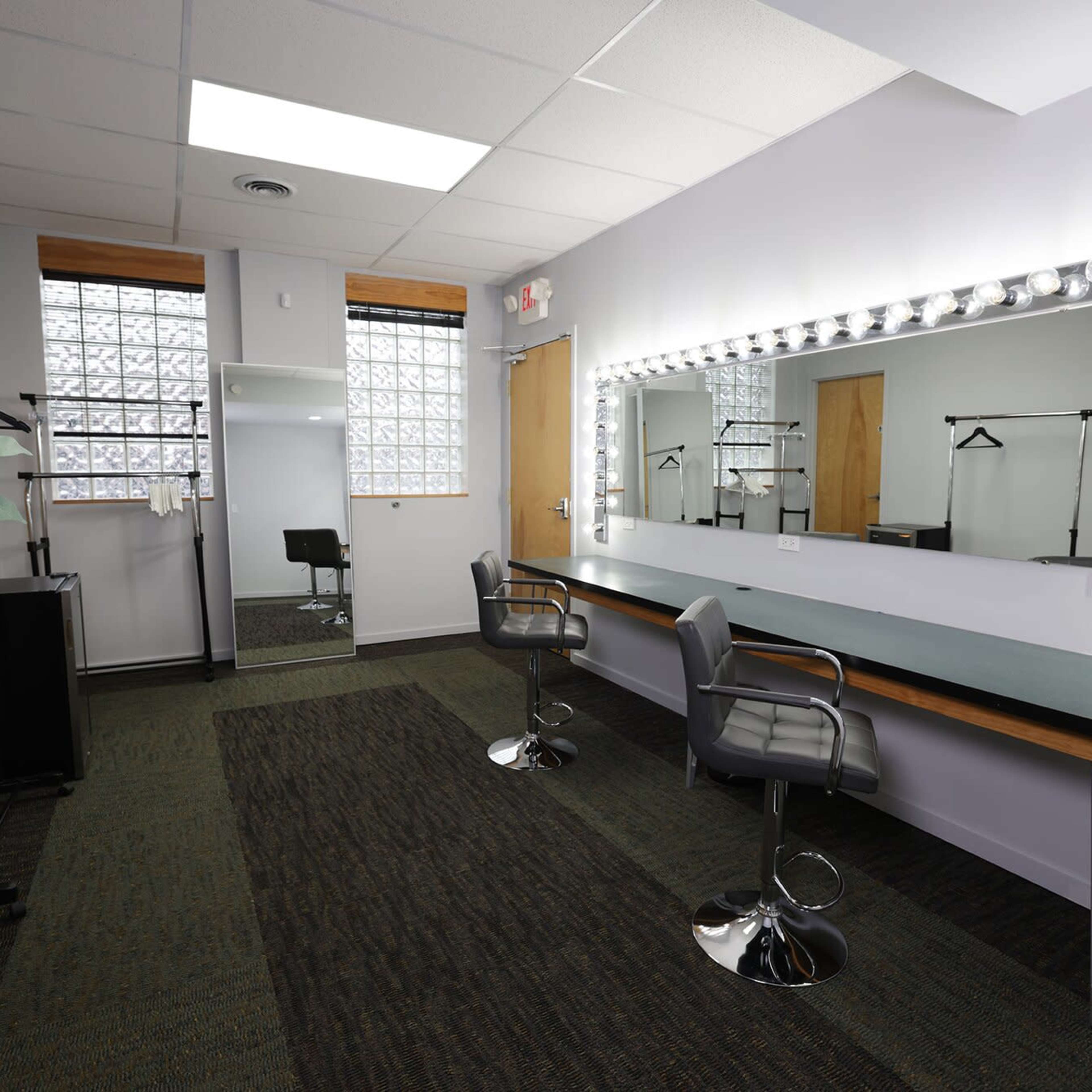 A dressing room with a large illuminated mirror, two barstools, and a green carpeted floor, along with a plant and clothing racks in the background.