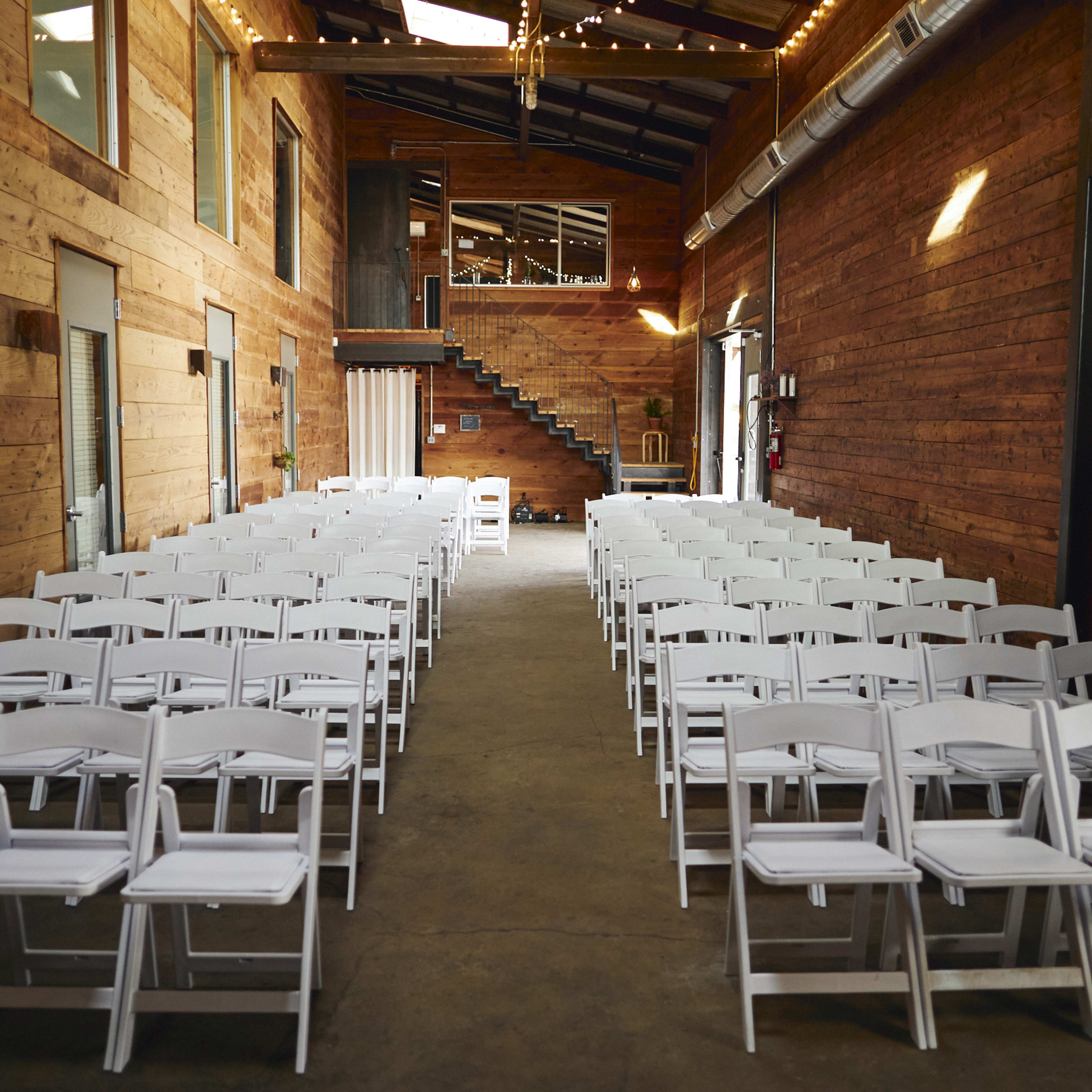 The interior of a rustic venue features rows of white folding chairs facing a wooden backdrop, with a staircase visible at the end of the room.