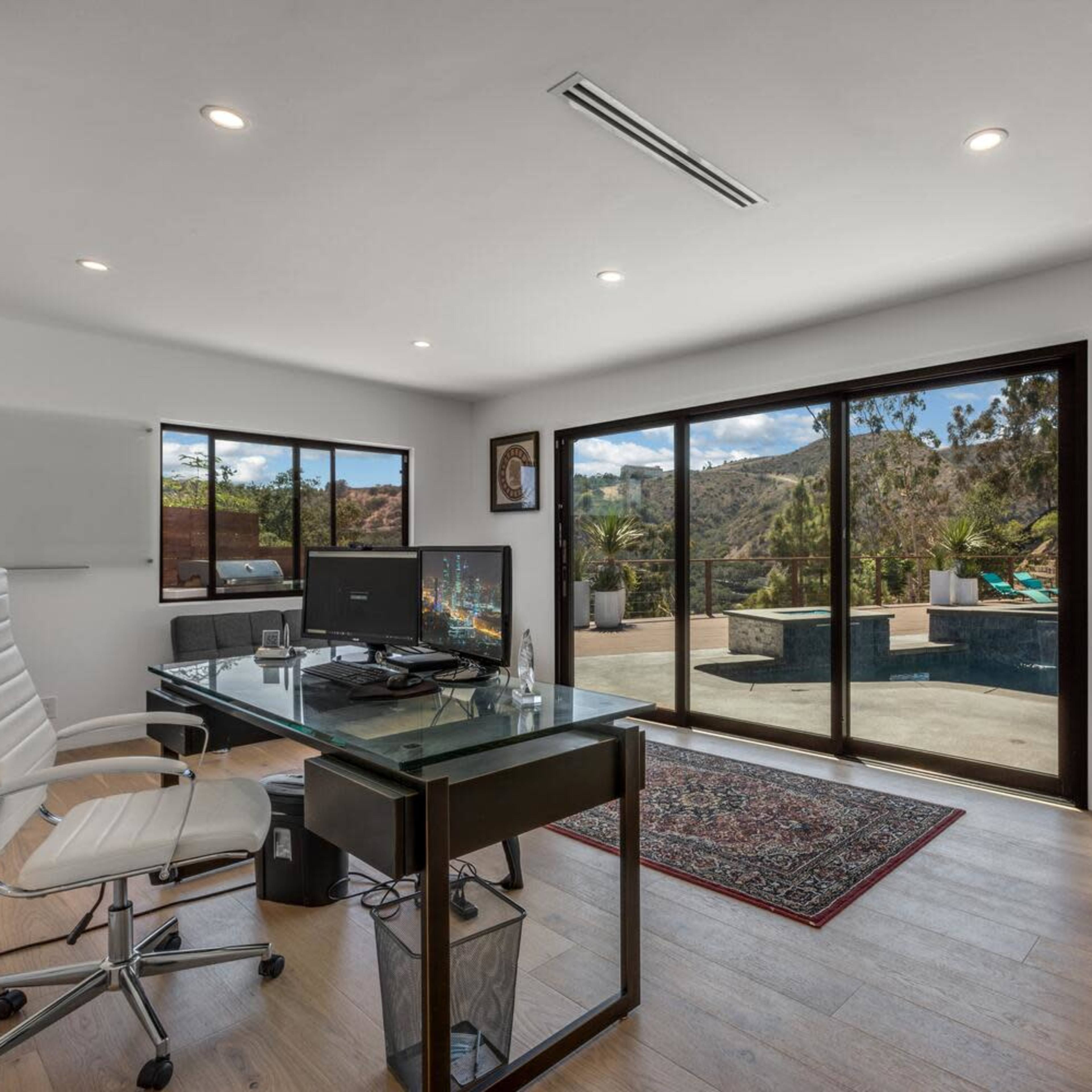 A modern office space features a glass desk with a computer, overlooking a patio and pool area through large sliding glass doors.