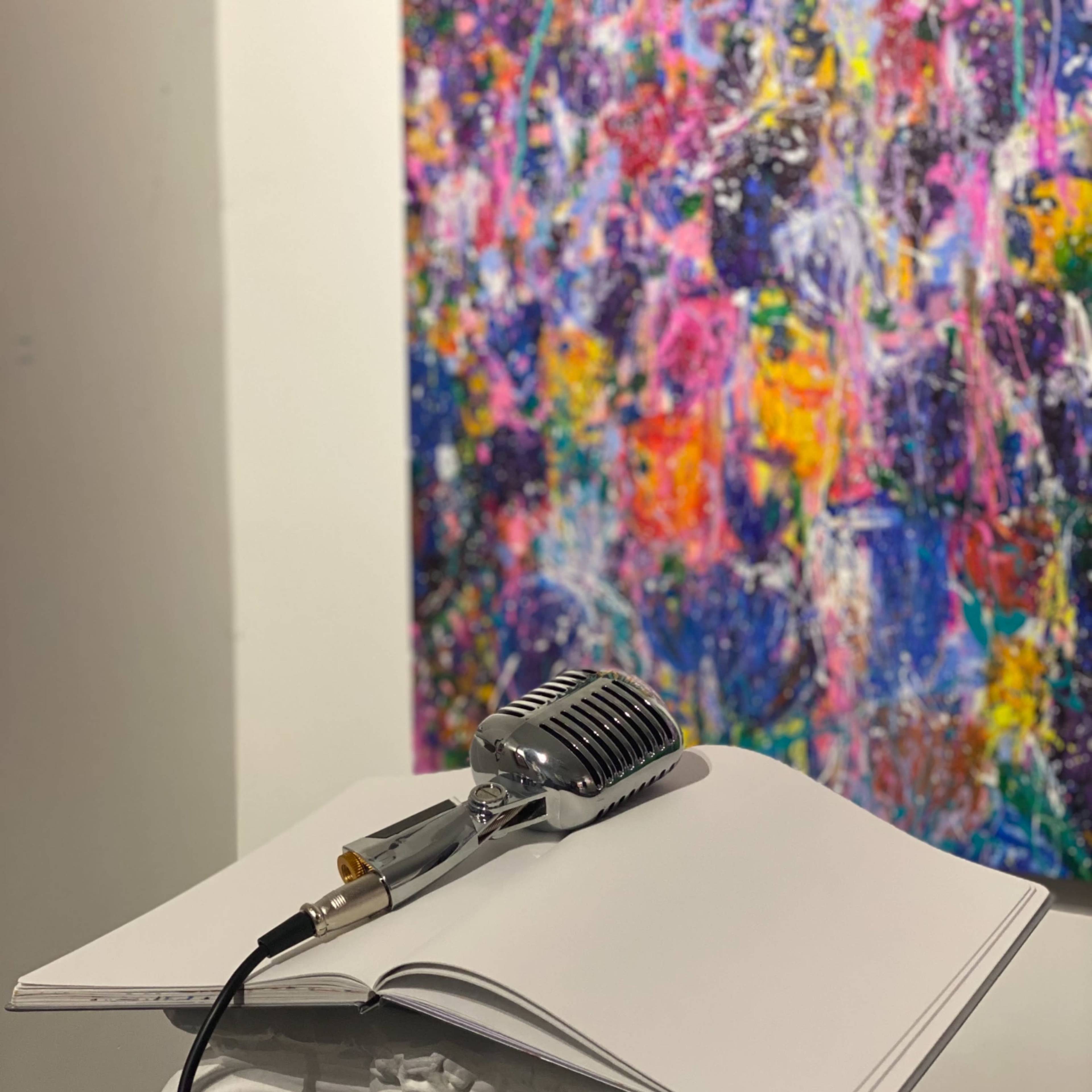 A vintage microphone sits on an open sketchbook placed on a decorative pedestal in front of a vibrant abstract painting.