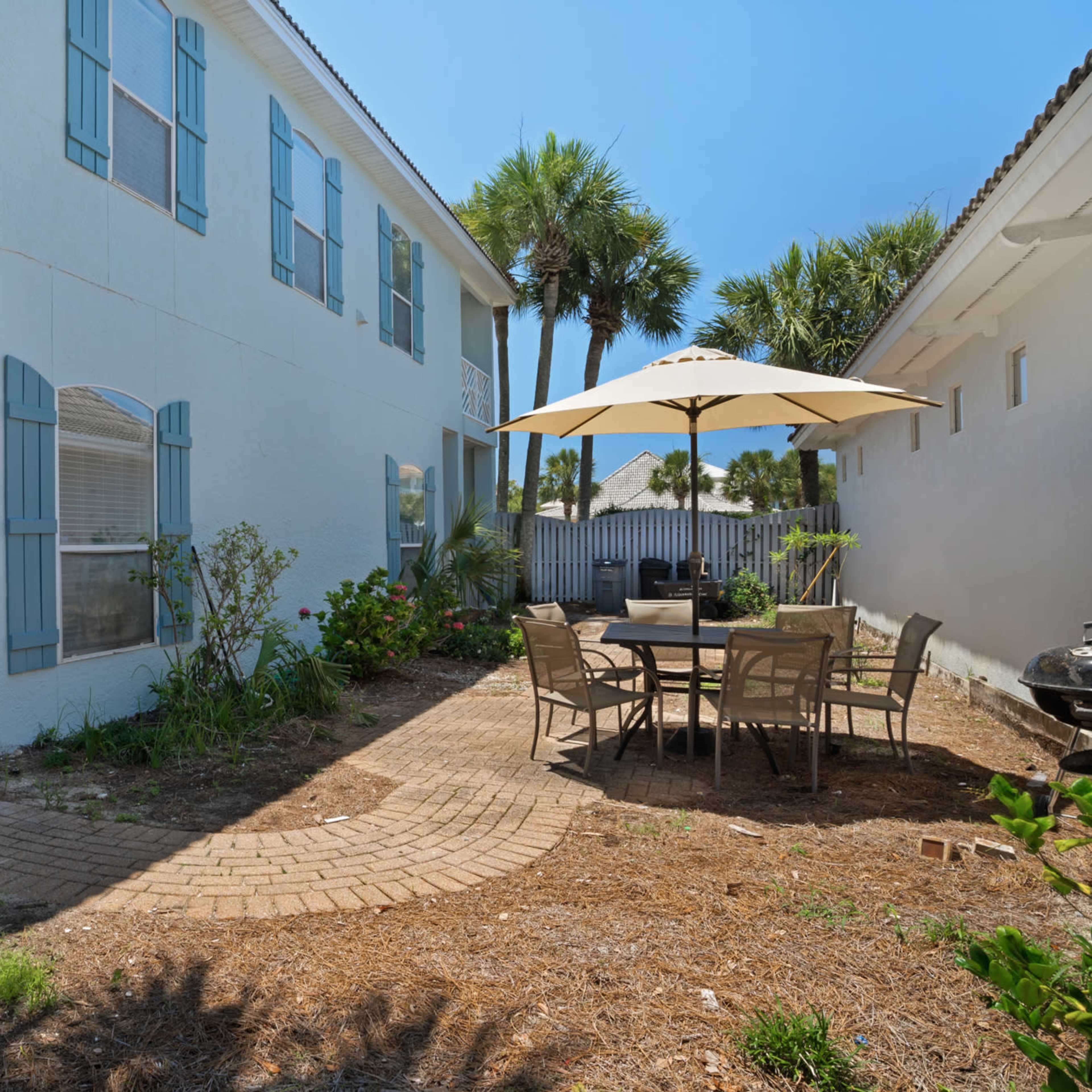 A brick path leads to a patio area with a table and chairs, surrounded by greenery next to a two-story building with blue shutters.