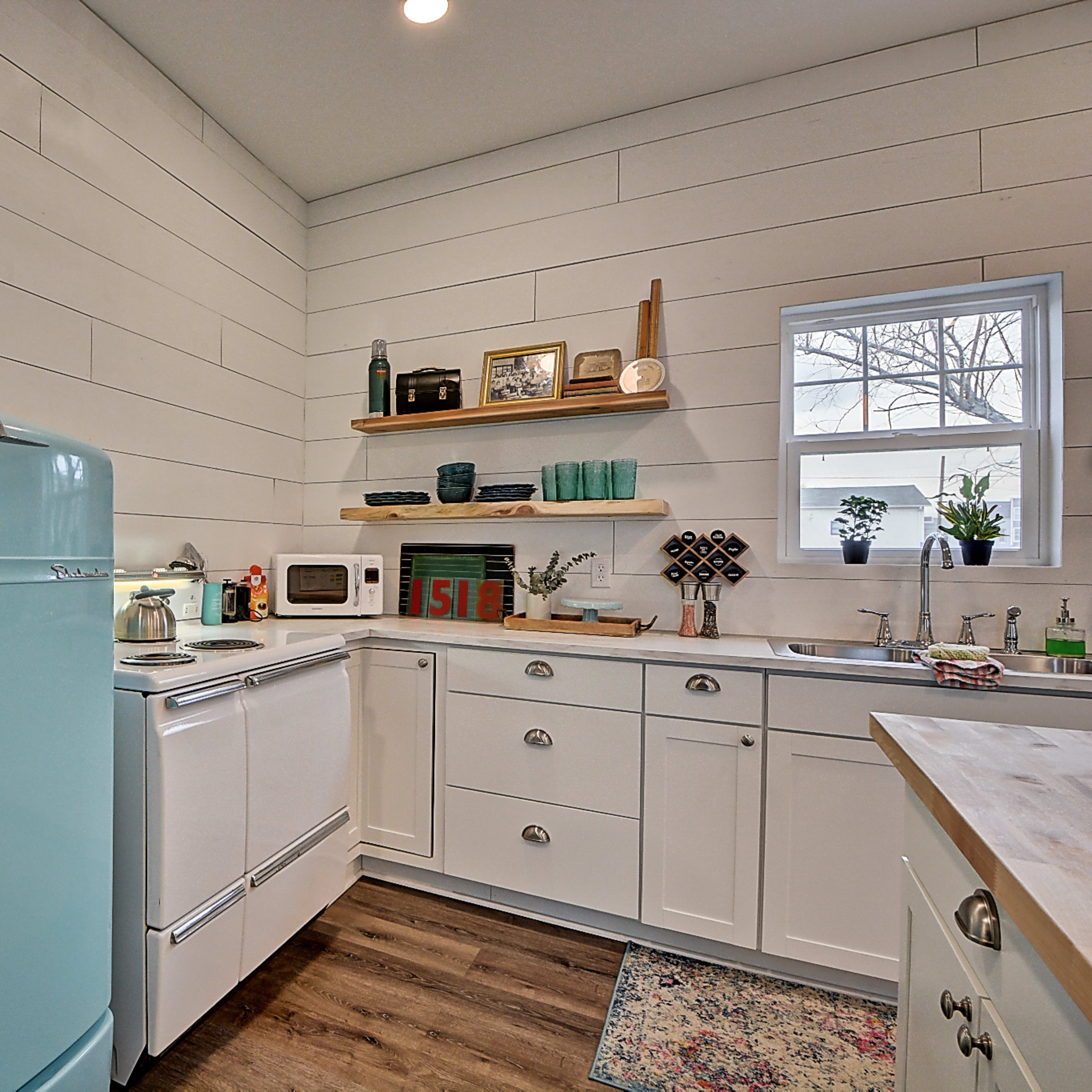 A bright kitchen features a retro turquoise refrigerator, white cabinets, and wood countertops, with a window providing natural light.