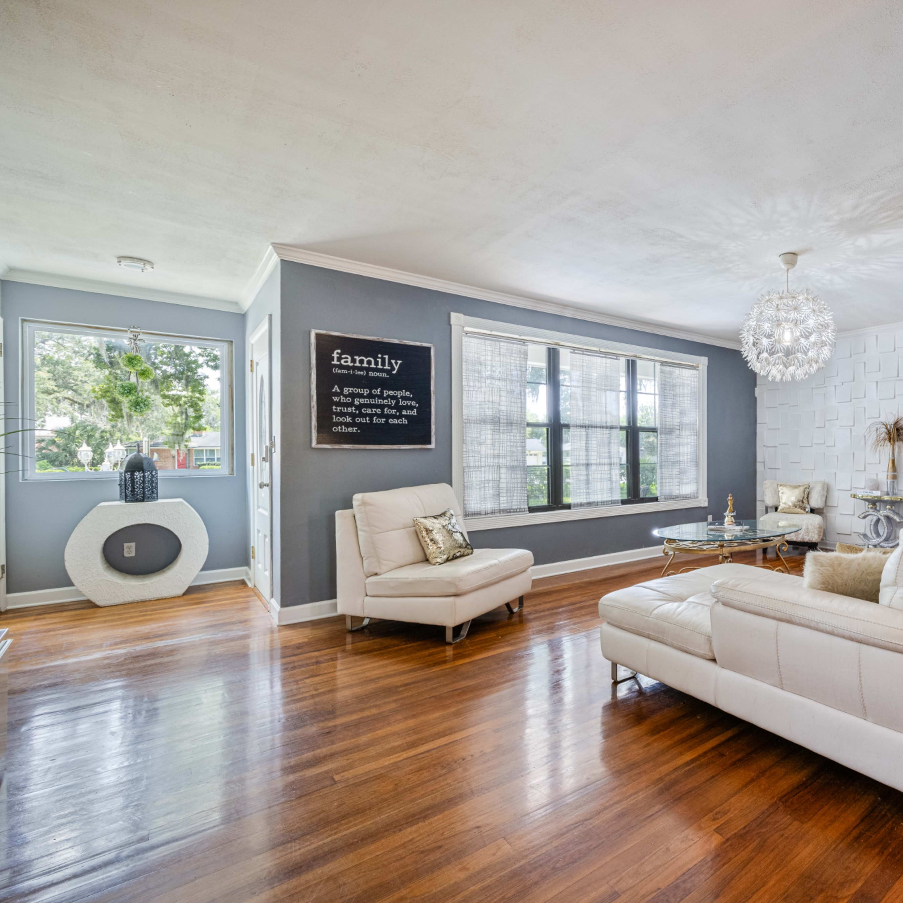 A modern living room features a white leather sofa, a glass coffee table, and a large window overlooking greenery, with a decorative wall art piece on the opposite side.