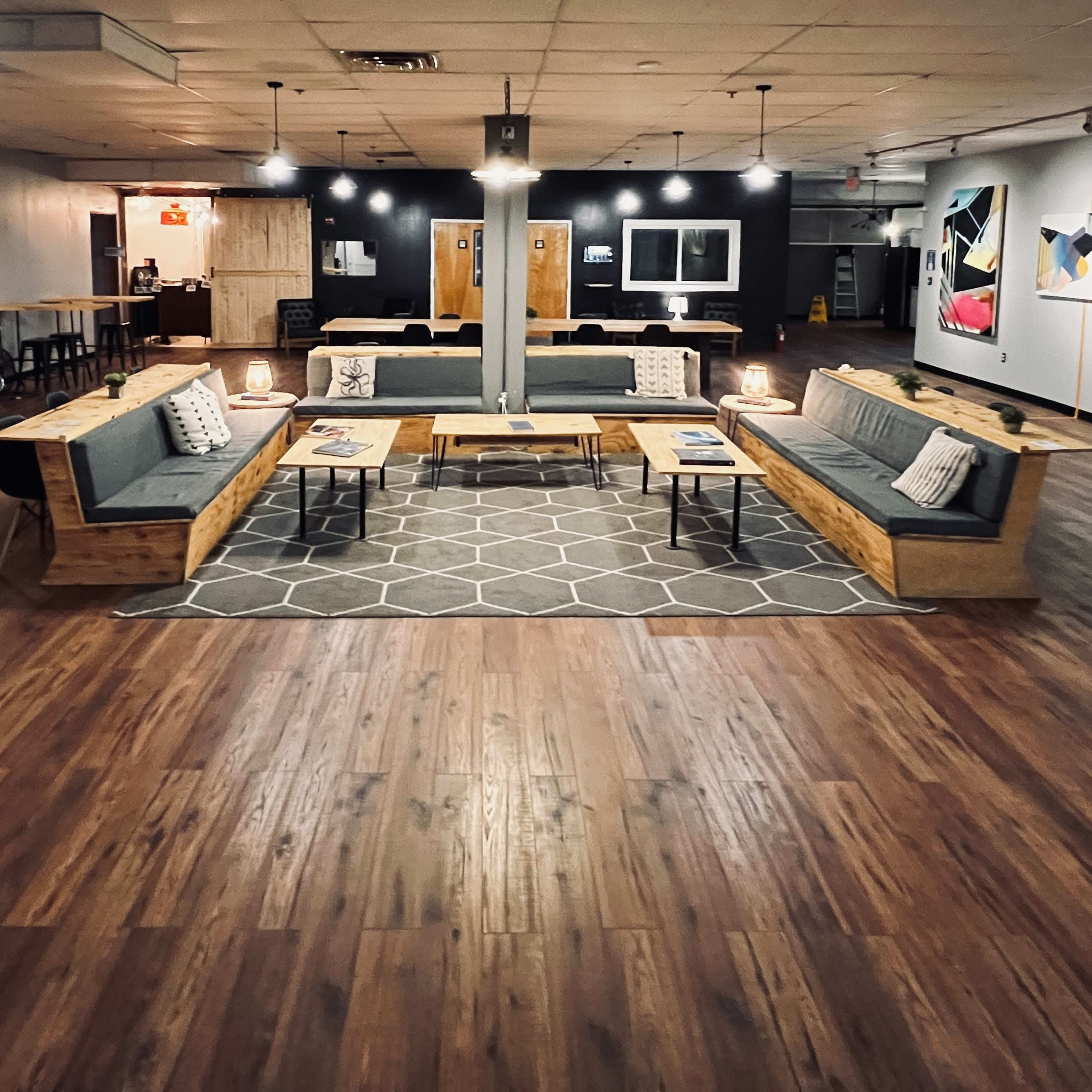 A spacious interior with wooden flooring, featuring a central seating area composed of modern sofas and tables, surrounded by framed artwork on the walls.
