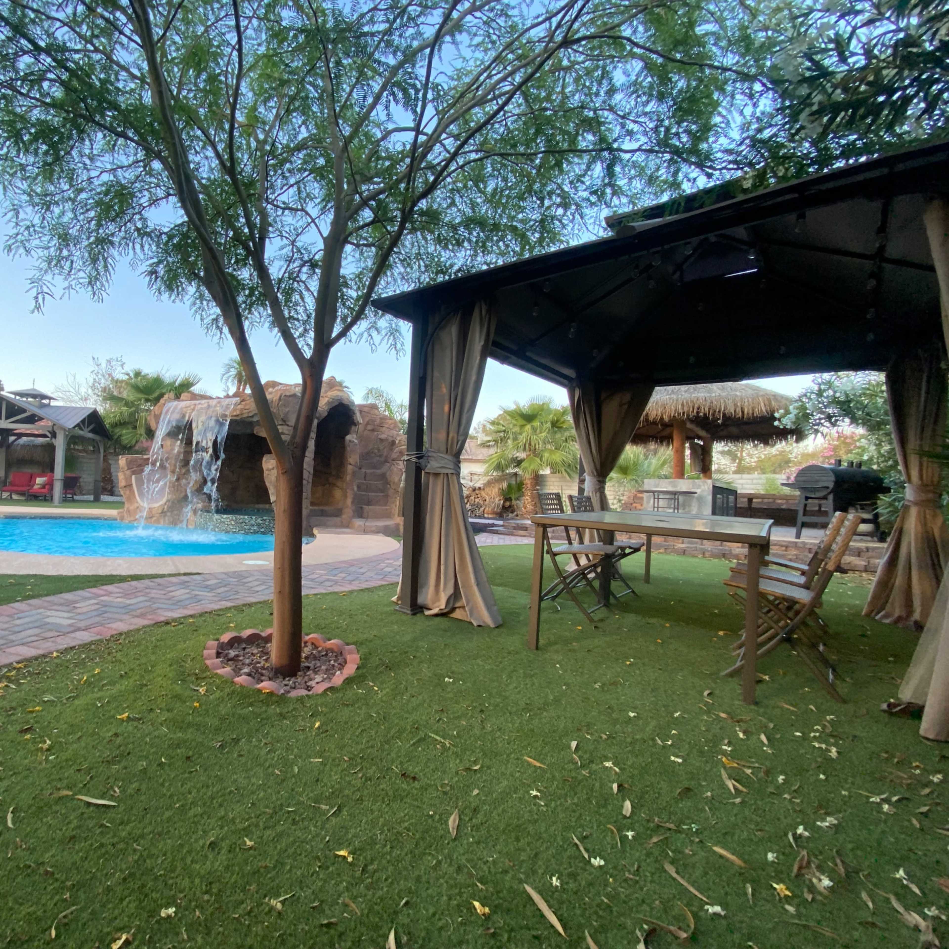 A landscaped backyard features a swimming pool with a waterfall, shaded seating areas, and a grassy lawn.