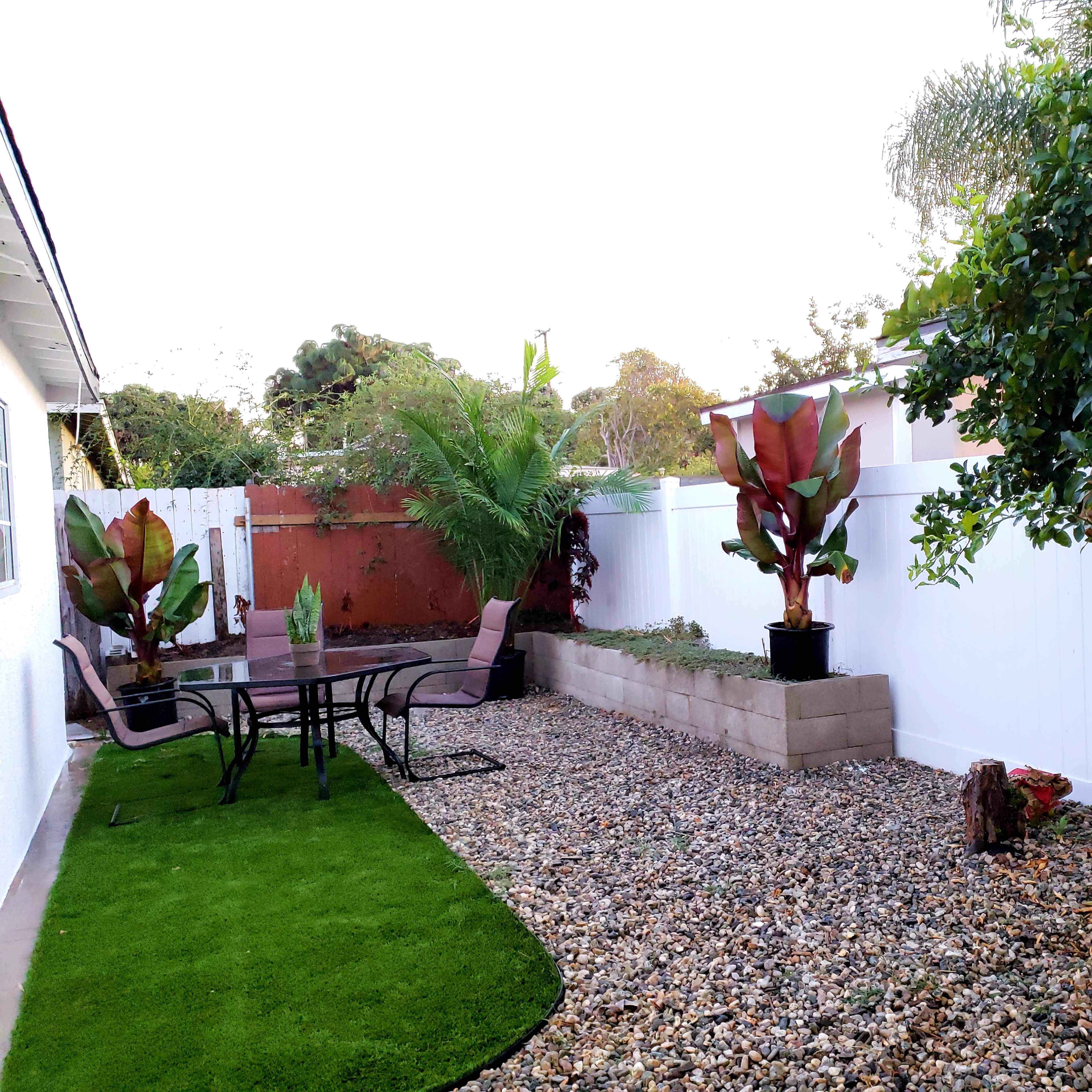 The backyard features a gravel section with a table and chairs, surrounded by potted plants and trees, alongside a small patch of artificial grass.