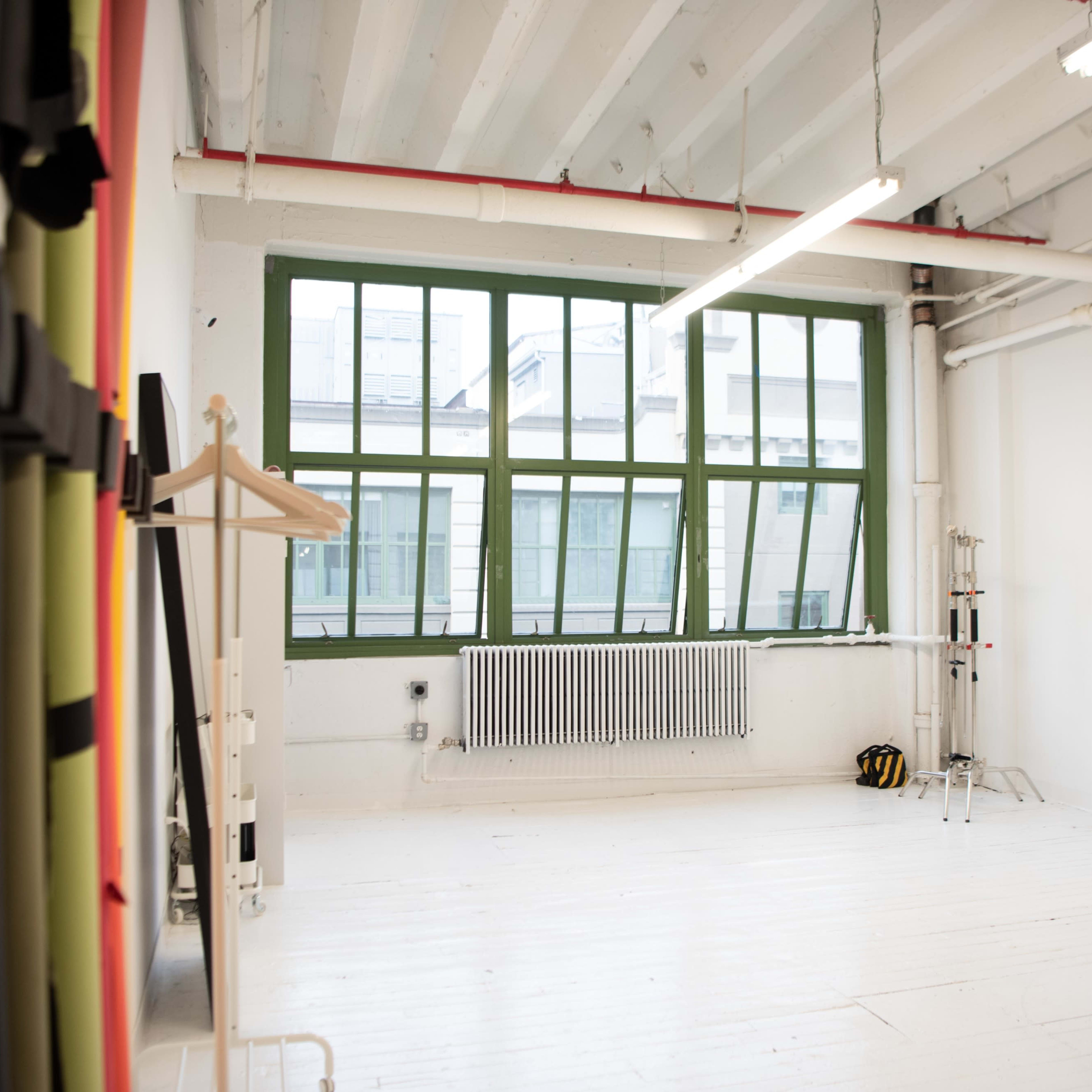 An empty studio space with large green-framed windows, white walls, wooden flooring, and minimalist furniture.