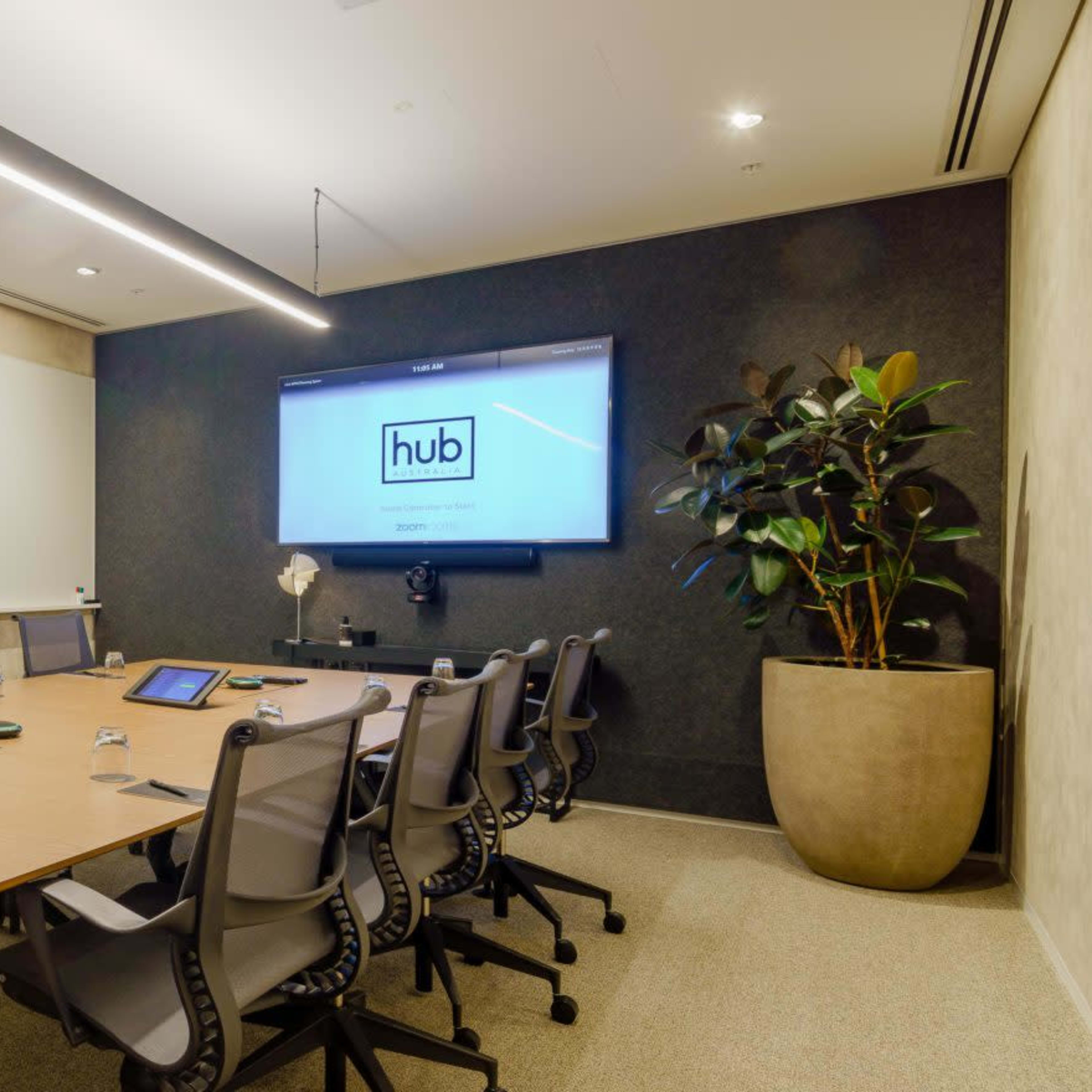 A modern conference room featuring a large table surrounded by office chairs, a wall-mounted screen, and a plant in a pot.