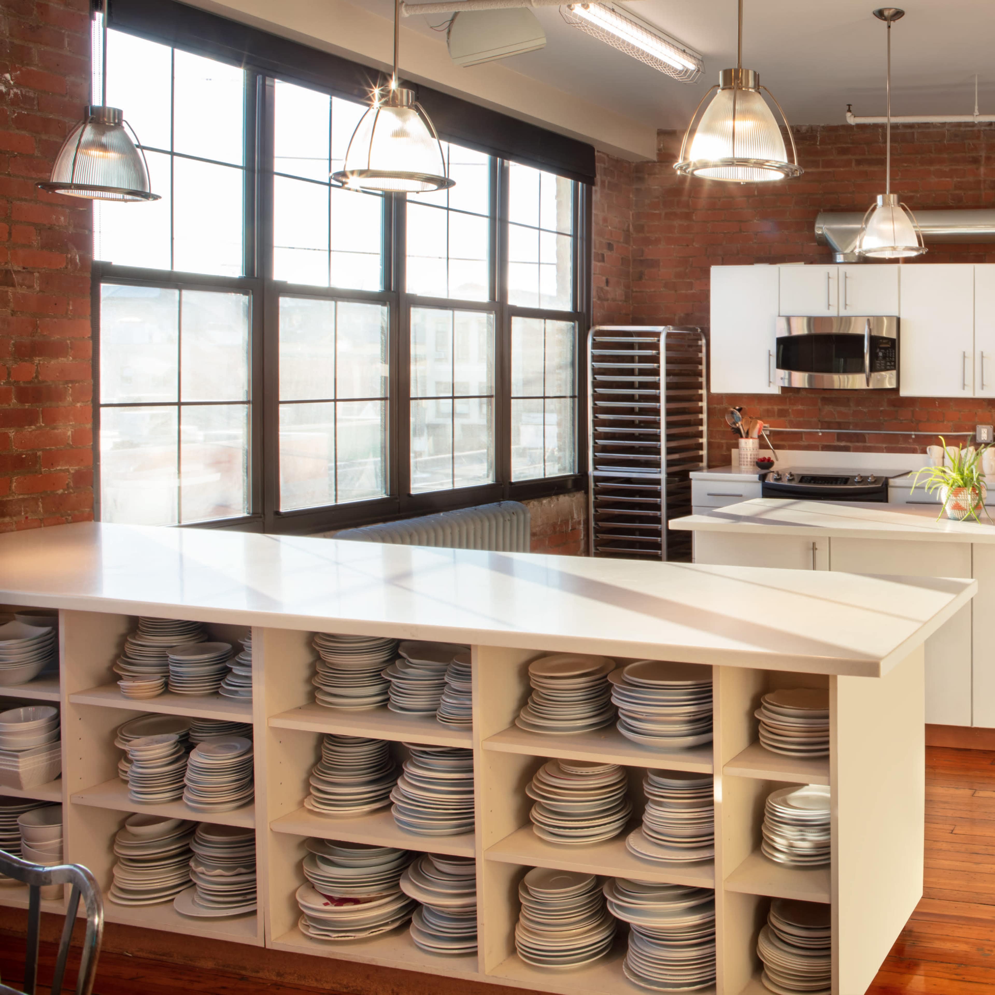 A modern kitchen with a long white island featuring open shelving filled with plates, stainless steel appliances, and large windows revealing a brick wall.