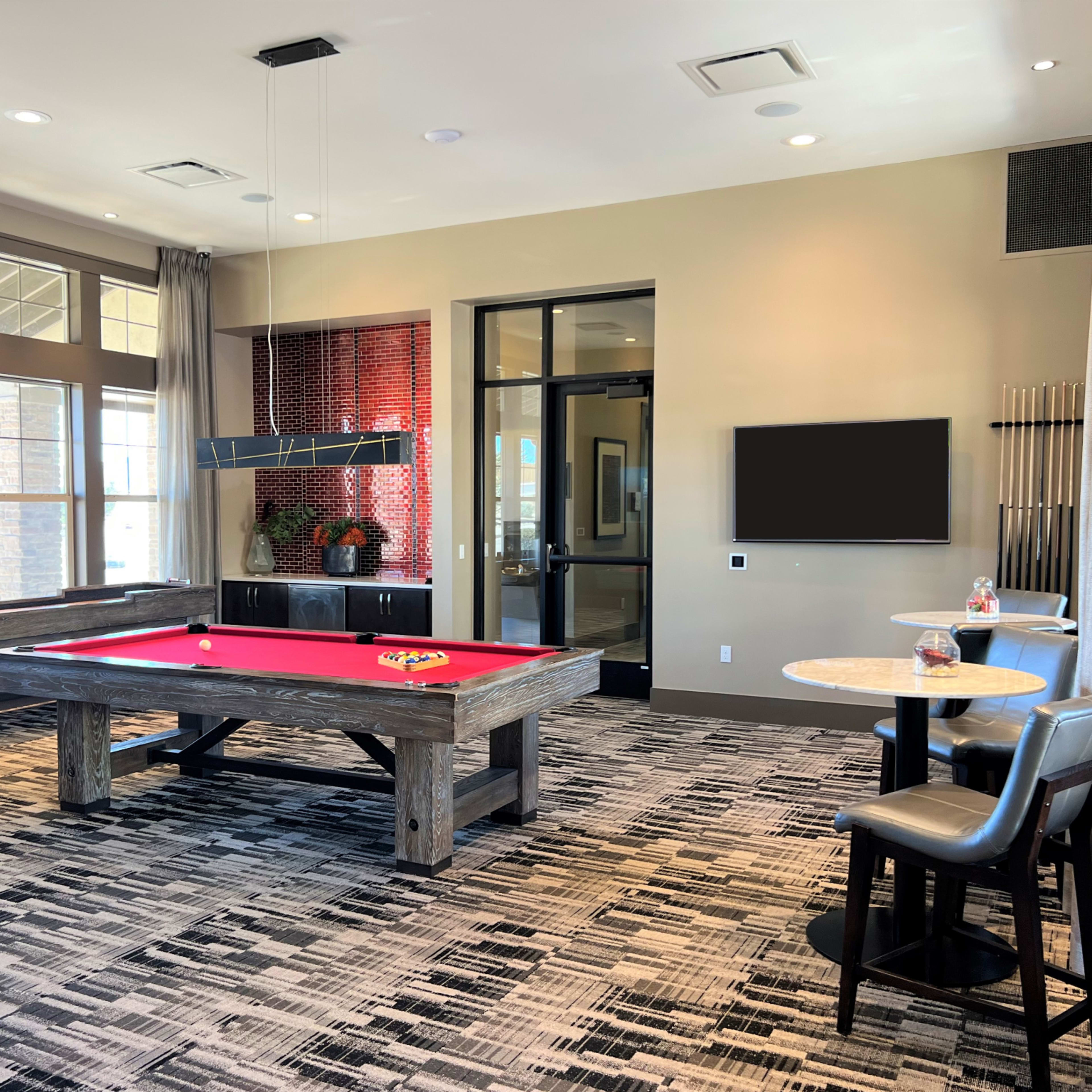 A billiard room with a red pool table, a seating area with two chairs and small round tables, and large windows allowing natural light.