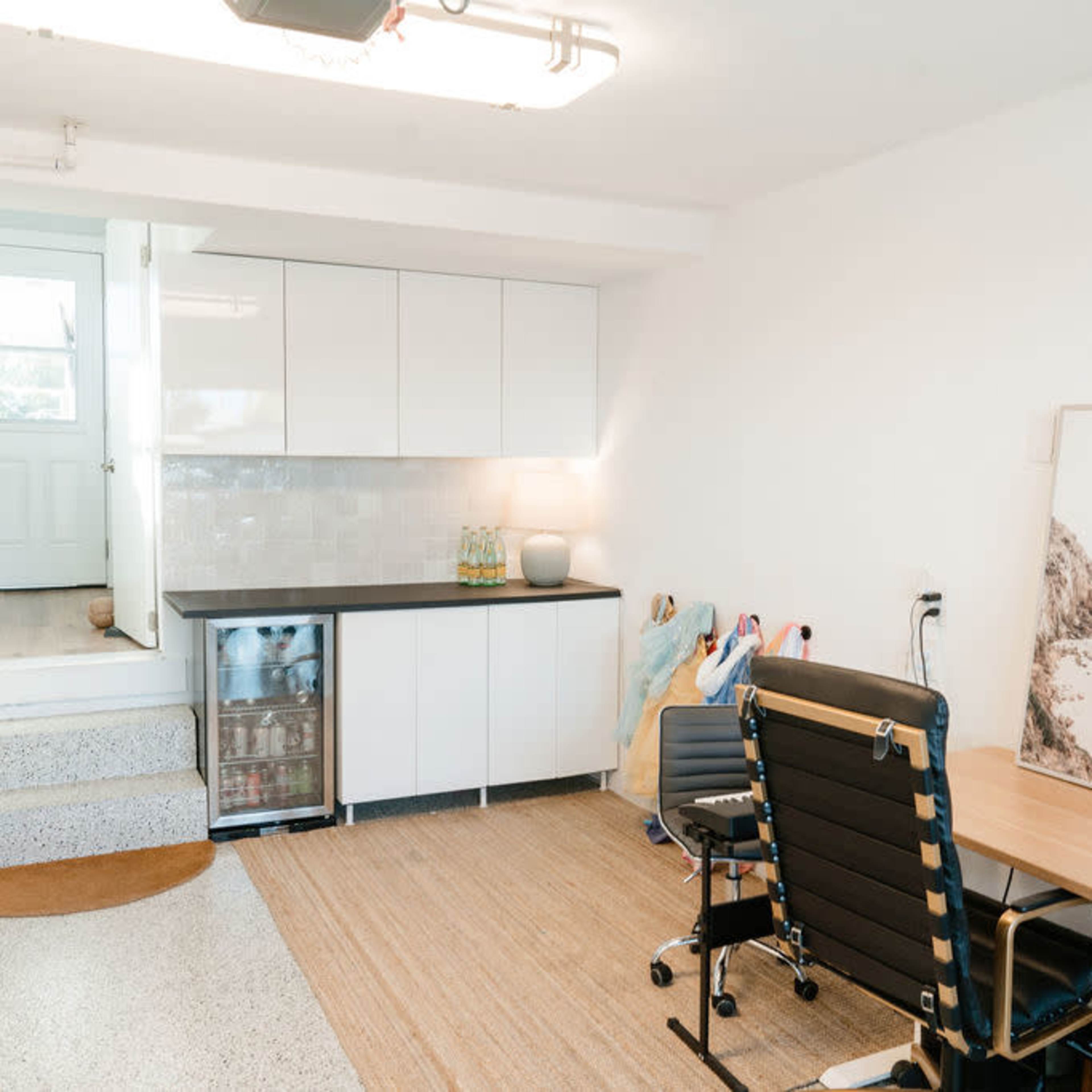 A clean, modern basement space featuring a kitchenette with cabinets and a mini-fridge, a desk with a chair, and a staircase leading to a door.