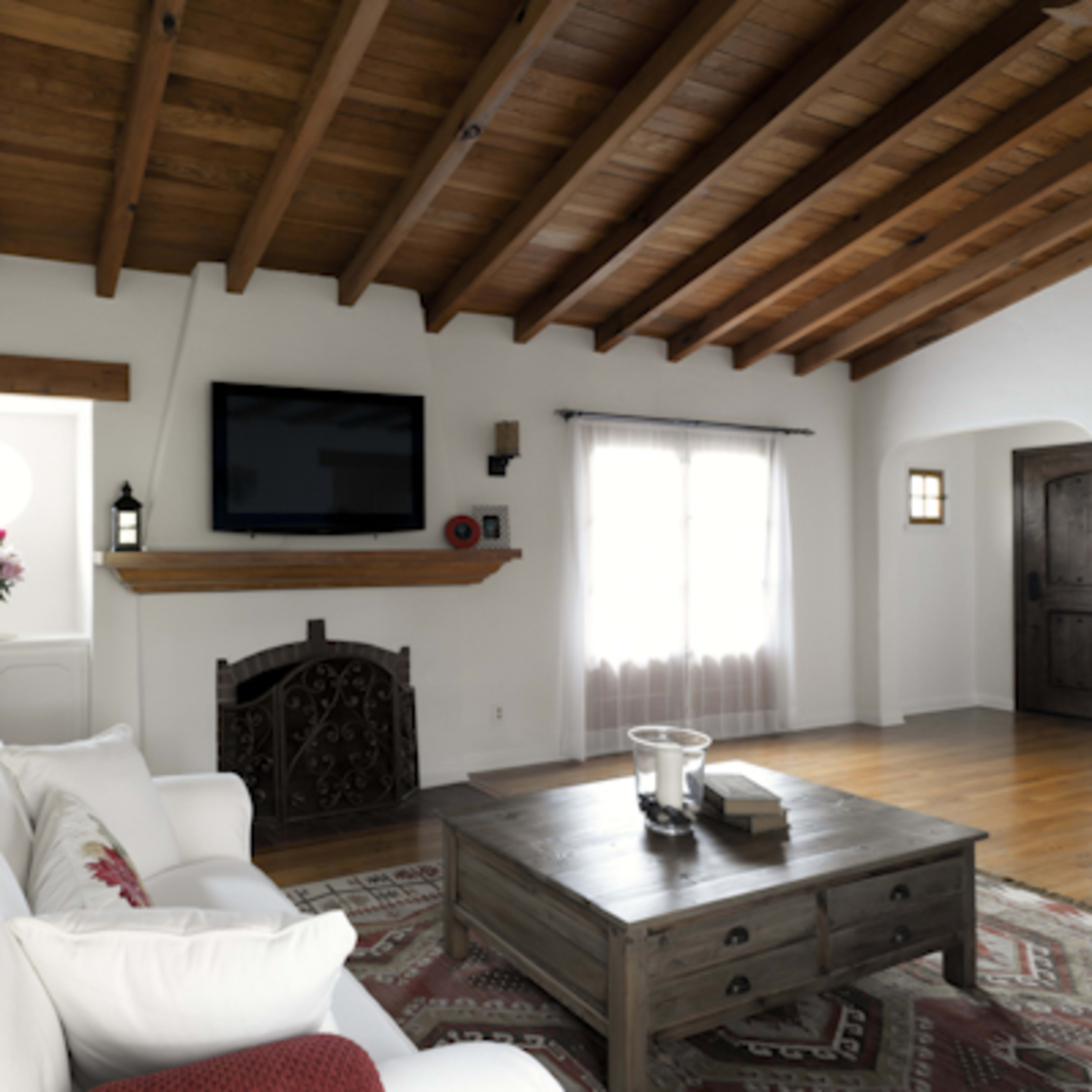 A living room with a white sofa, a wooden coffee table, a flat-screen TV mounted above a wooden mantel, and large windows with sheer curtains.