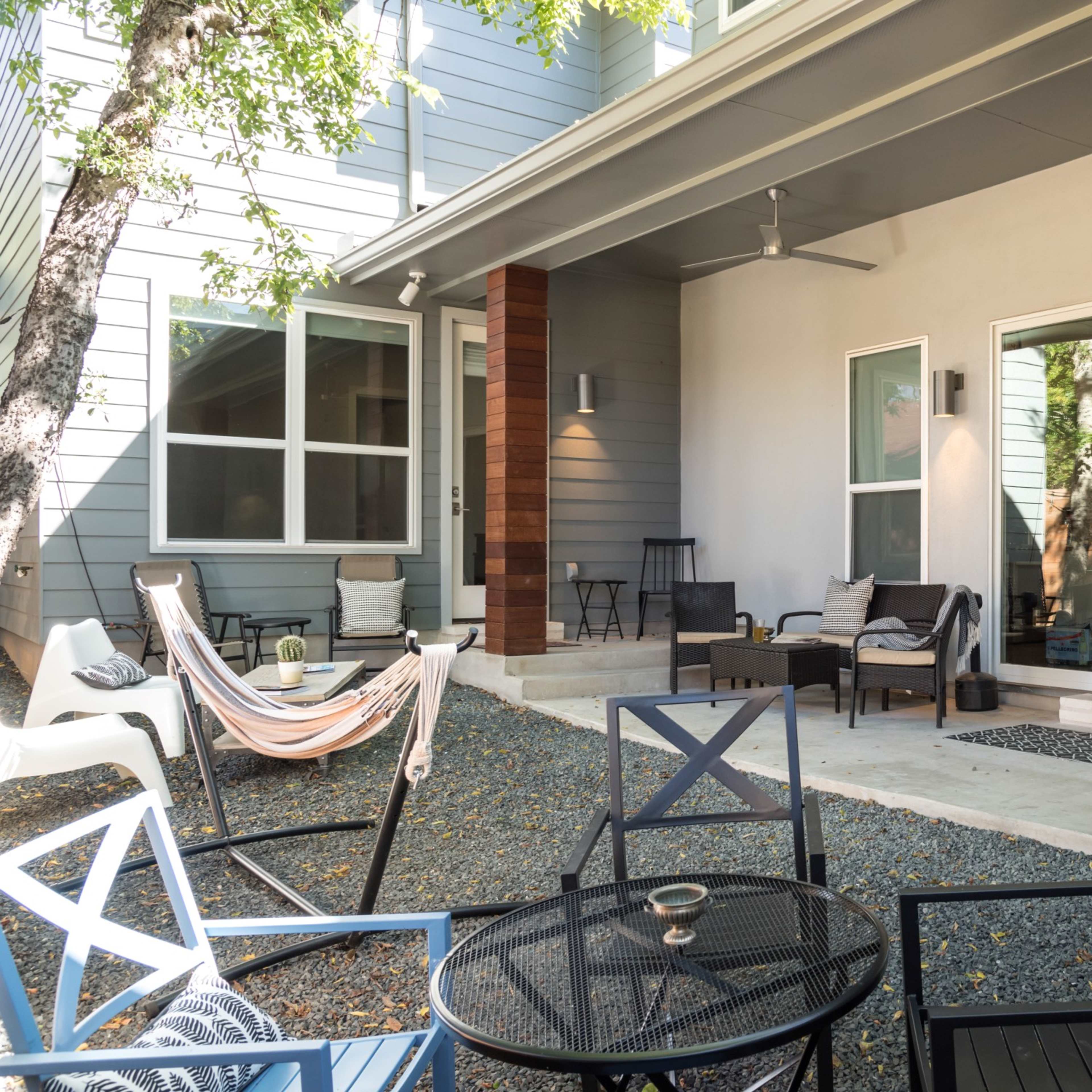 A patio area with outdoor seating, a hammock, and gravel flooring, surrounded by trees and sliding glass doors leading inside.