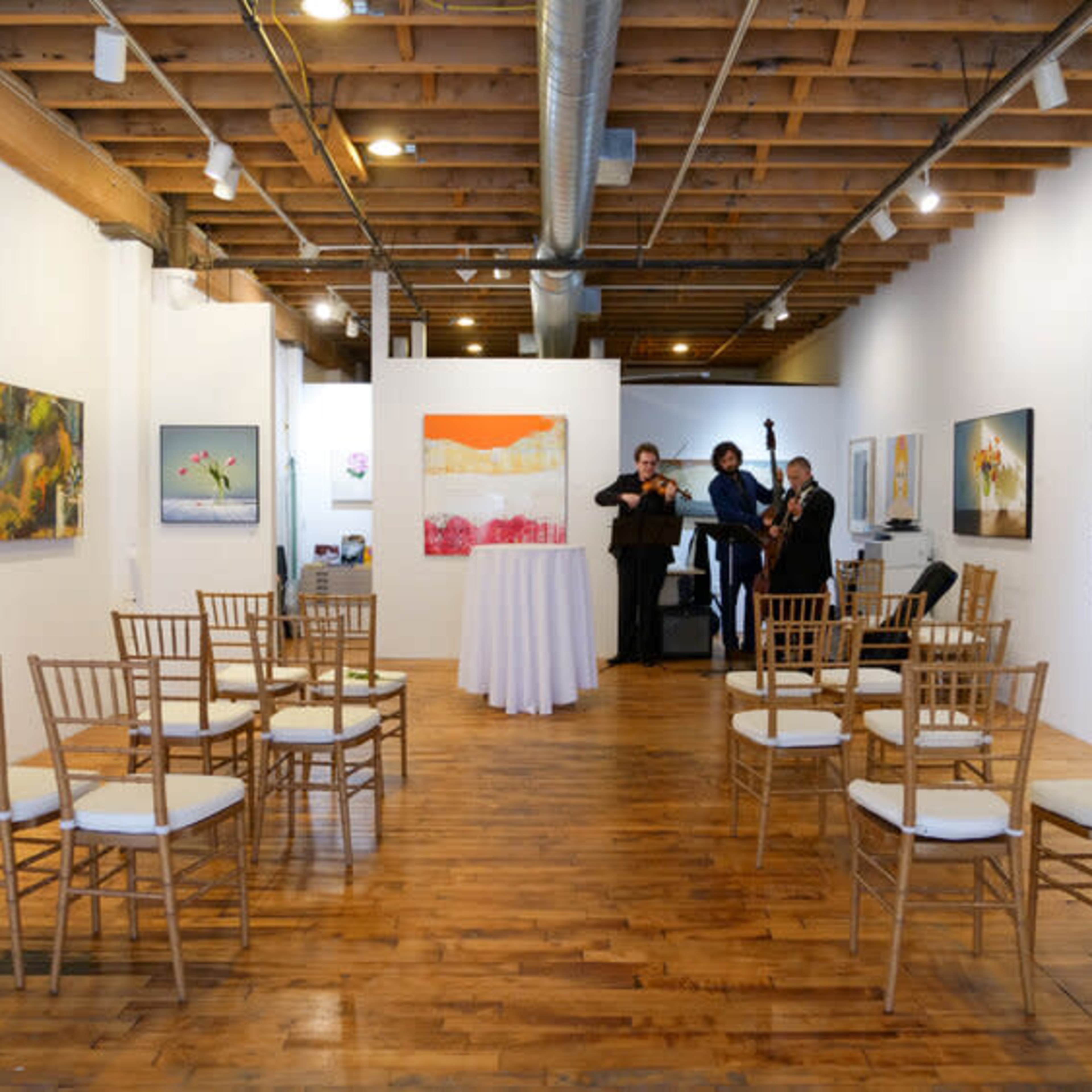 A spacious art gallery features wooden flooring and several chairs arranged in two semi-circles, with musicians performing near colorful abstract paintings.