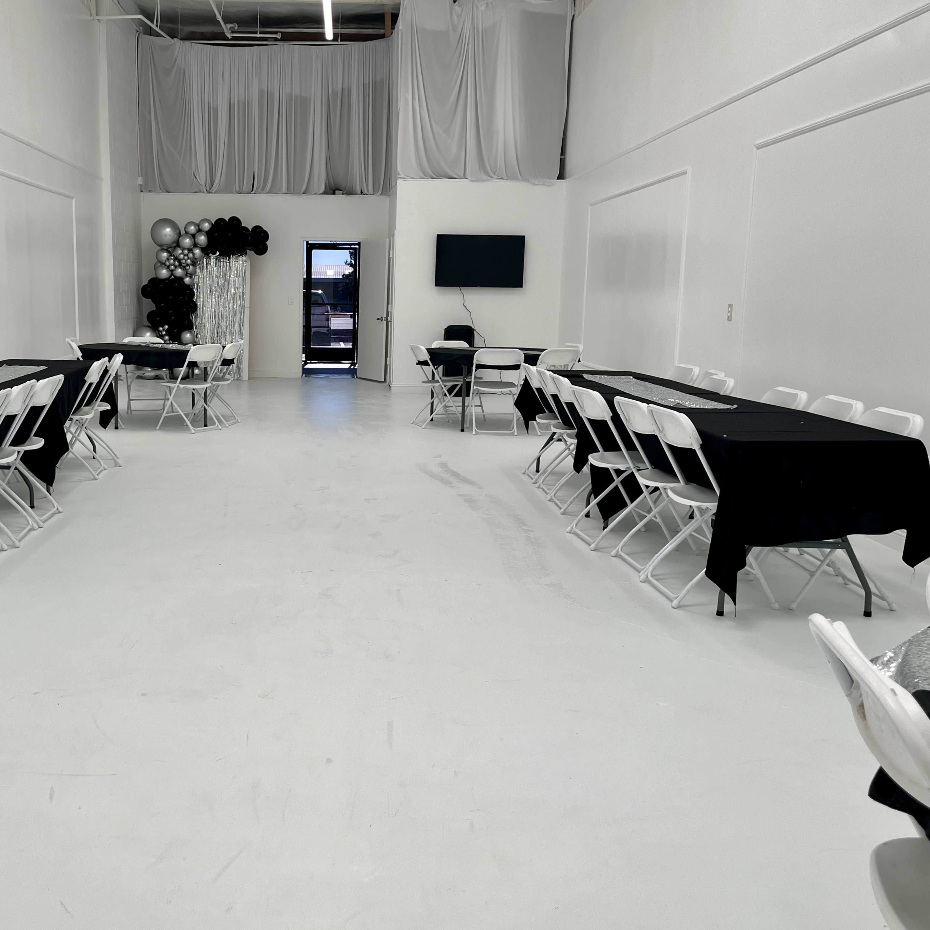 A spacious, empty room set up with black tablecloths over white folding tables arranged in two rows, with a black and white decorative arrangement in the corner.