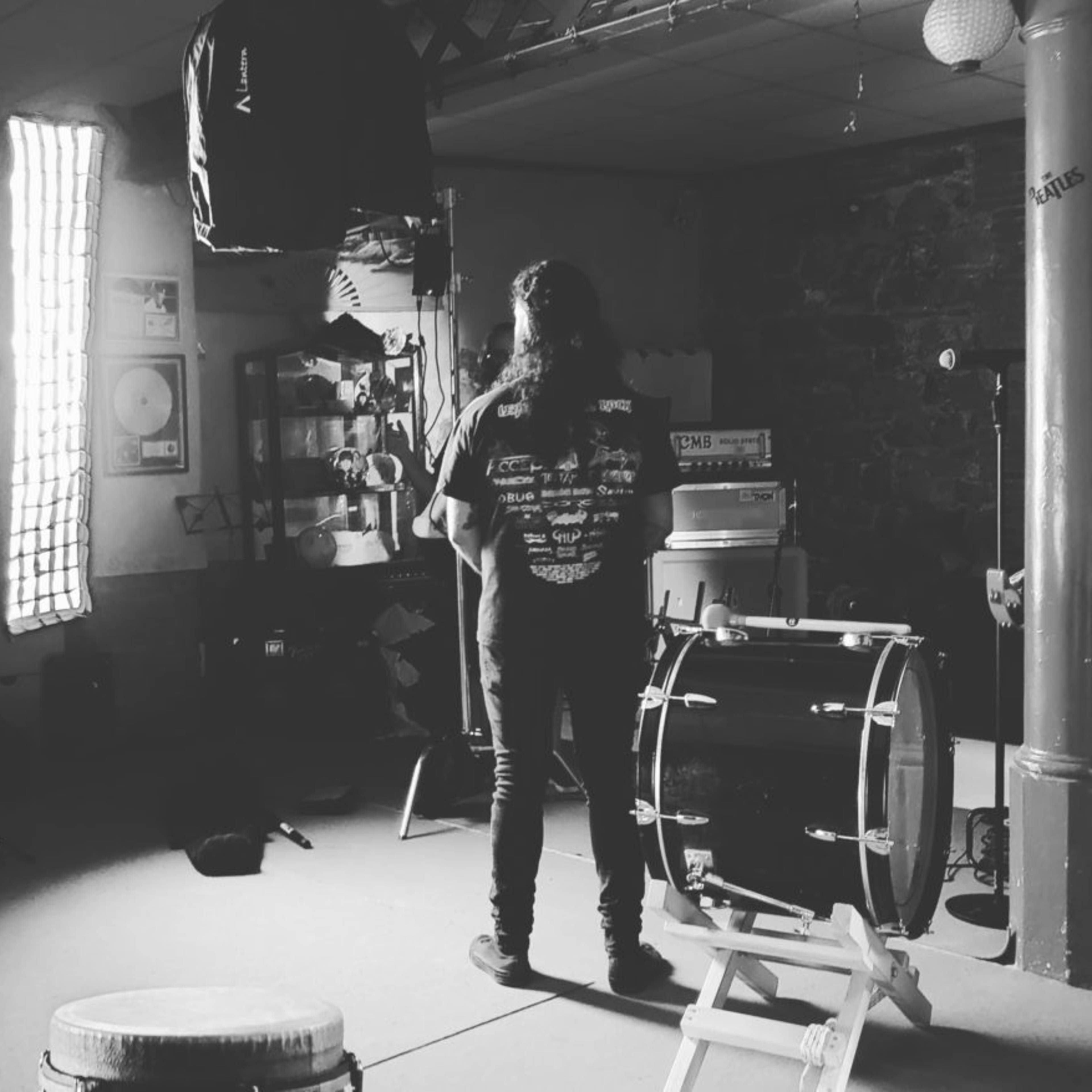 A person stands in a dimly lit room with instruments and music equipment while another individual is taking a photo.