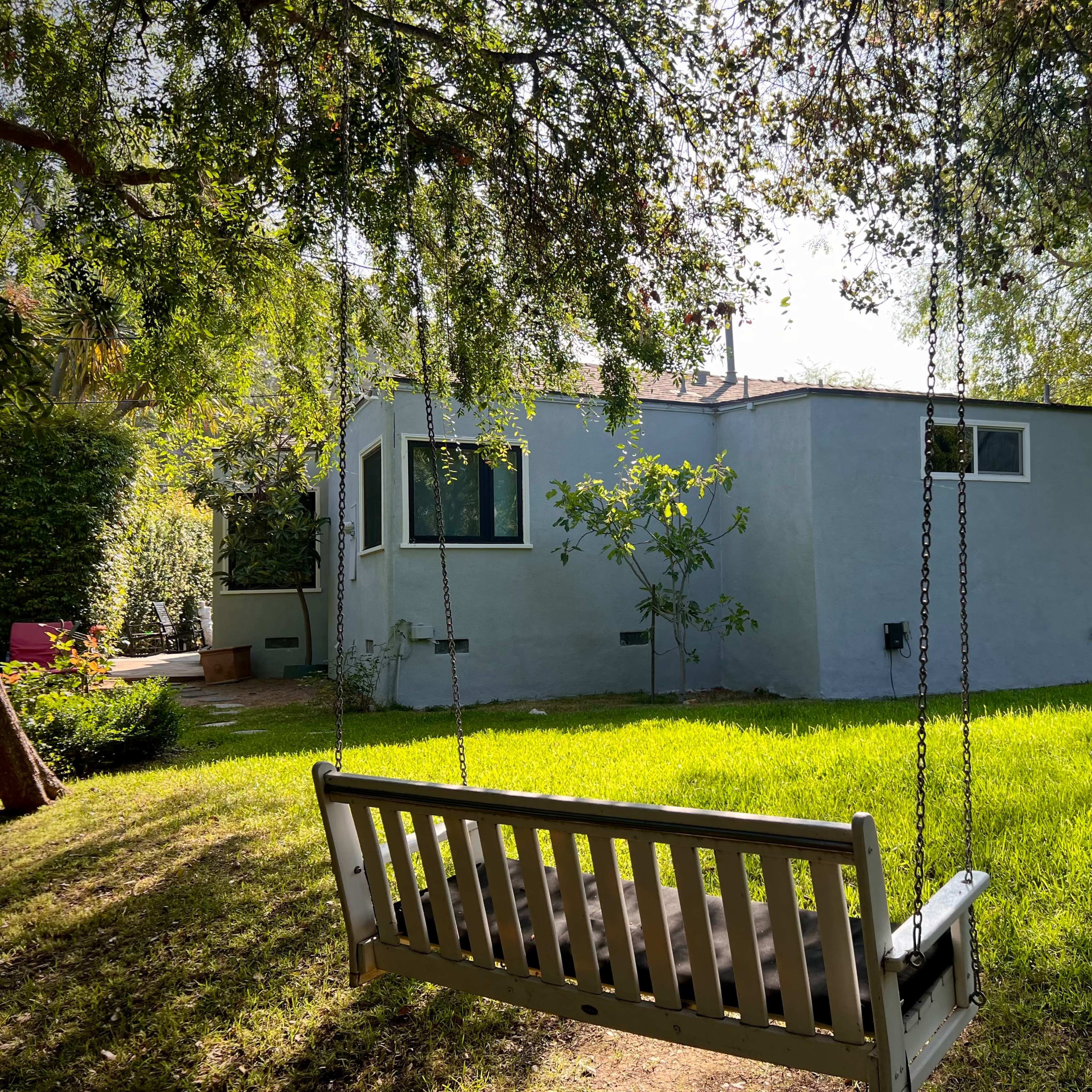 A wooden swing hangs in the foreground of a backyard, facing a pale gray house surrounded by lush greenery.