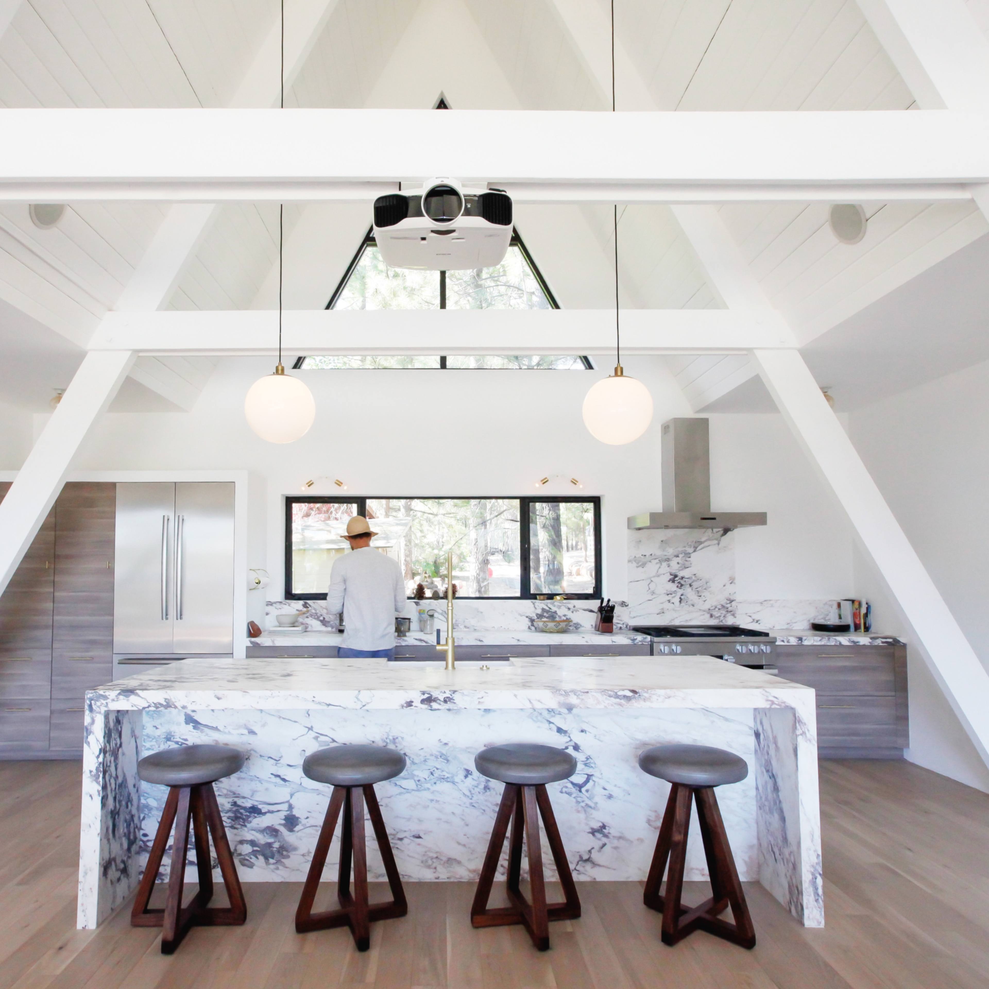 A modern kitchen with a marble island and four wooden stools, featuring high ceilings and large windows that provide natural light.