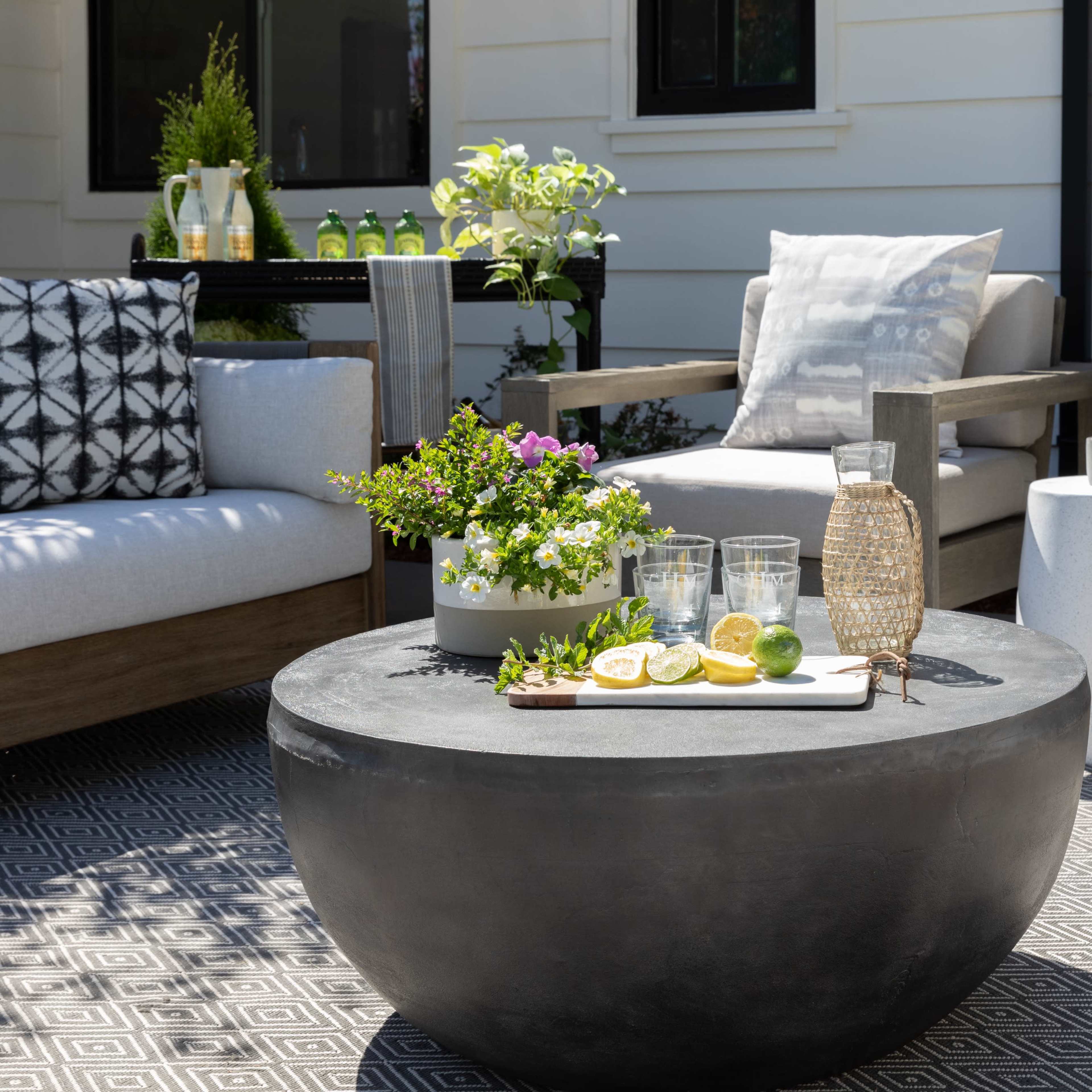 A modern outdoor seating area features two chairs with decorative cushions, a round black table topped with a tray of drinks and fresh fruit, and potted plants in the background.